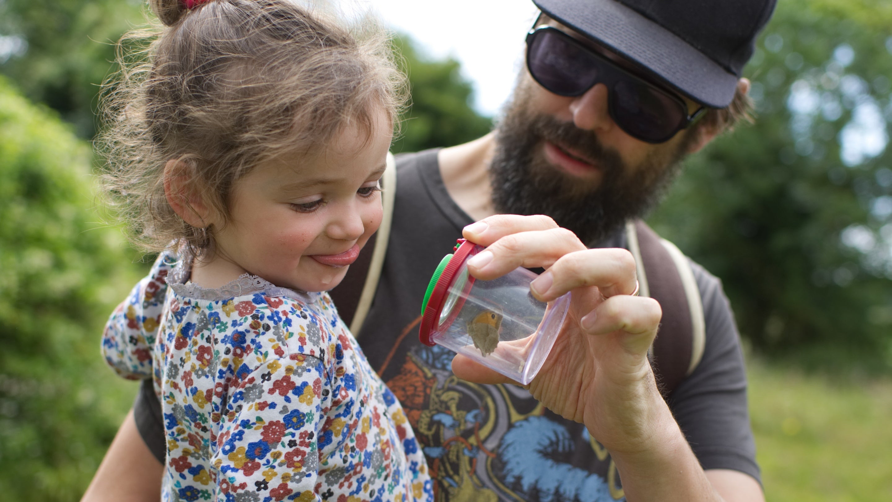 A father holds his daughter in his arms as they look at a transparent pot containing a butterfly