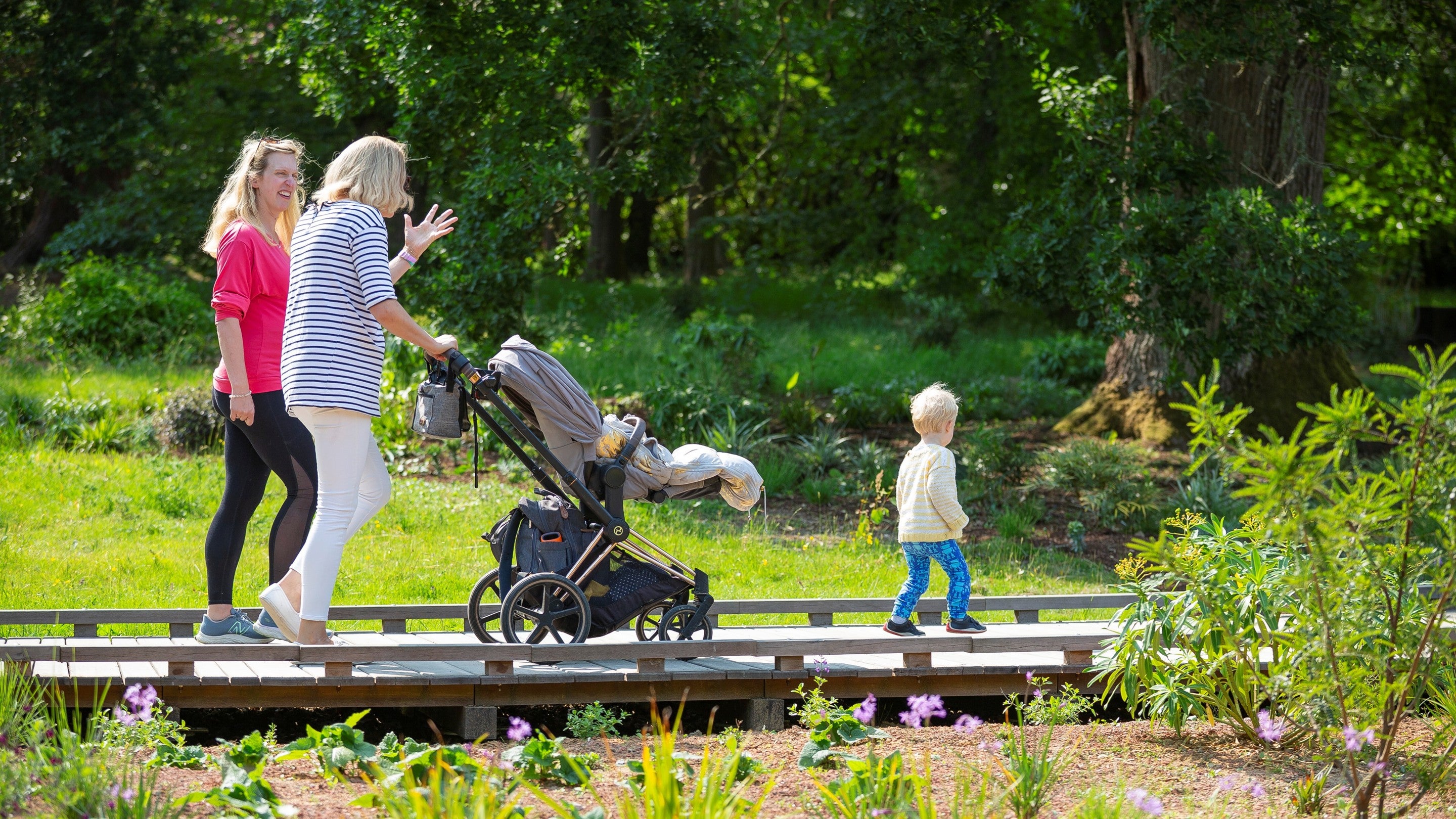 Two adults walk across a wooden path in a landscaped garden, pushing a stroller while a small child walks ahead. The path is surrounded by green grass, planting beds, and trees in bright daylight.