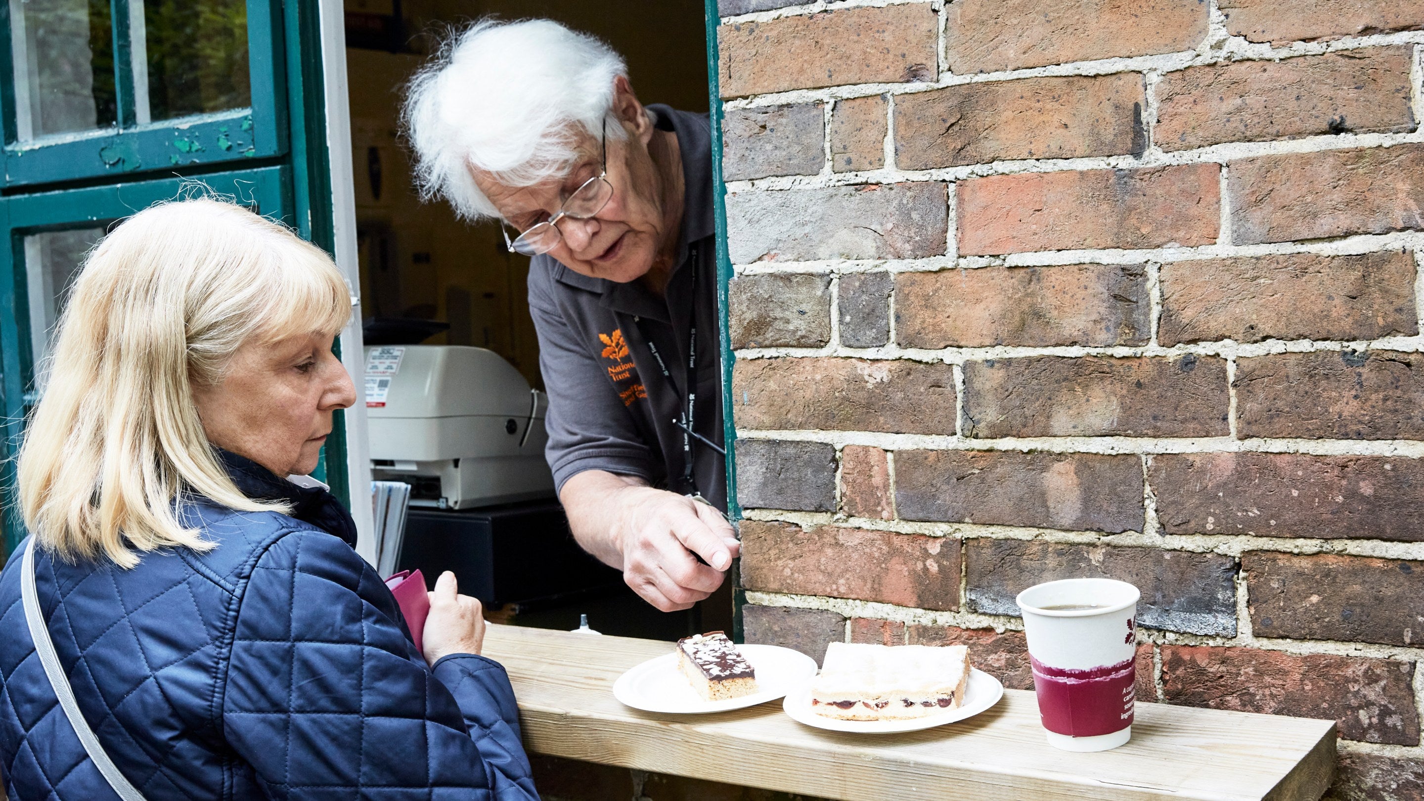 A volunteer sells a cake, sandwich and hot drink to a visitor, through the window, at Sheffield Park and Garden, East Sussex