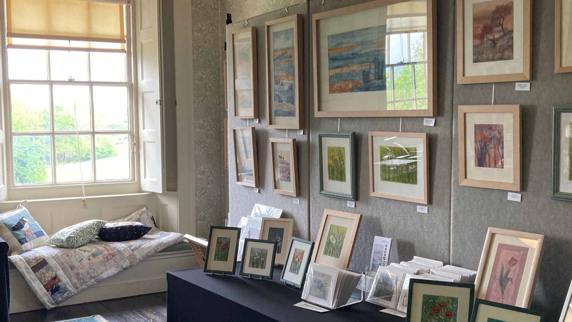 Paintings and pictures exhibited for sale in a room with a large window overlooking greenery.