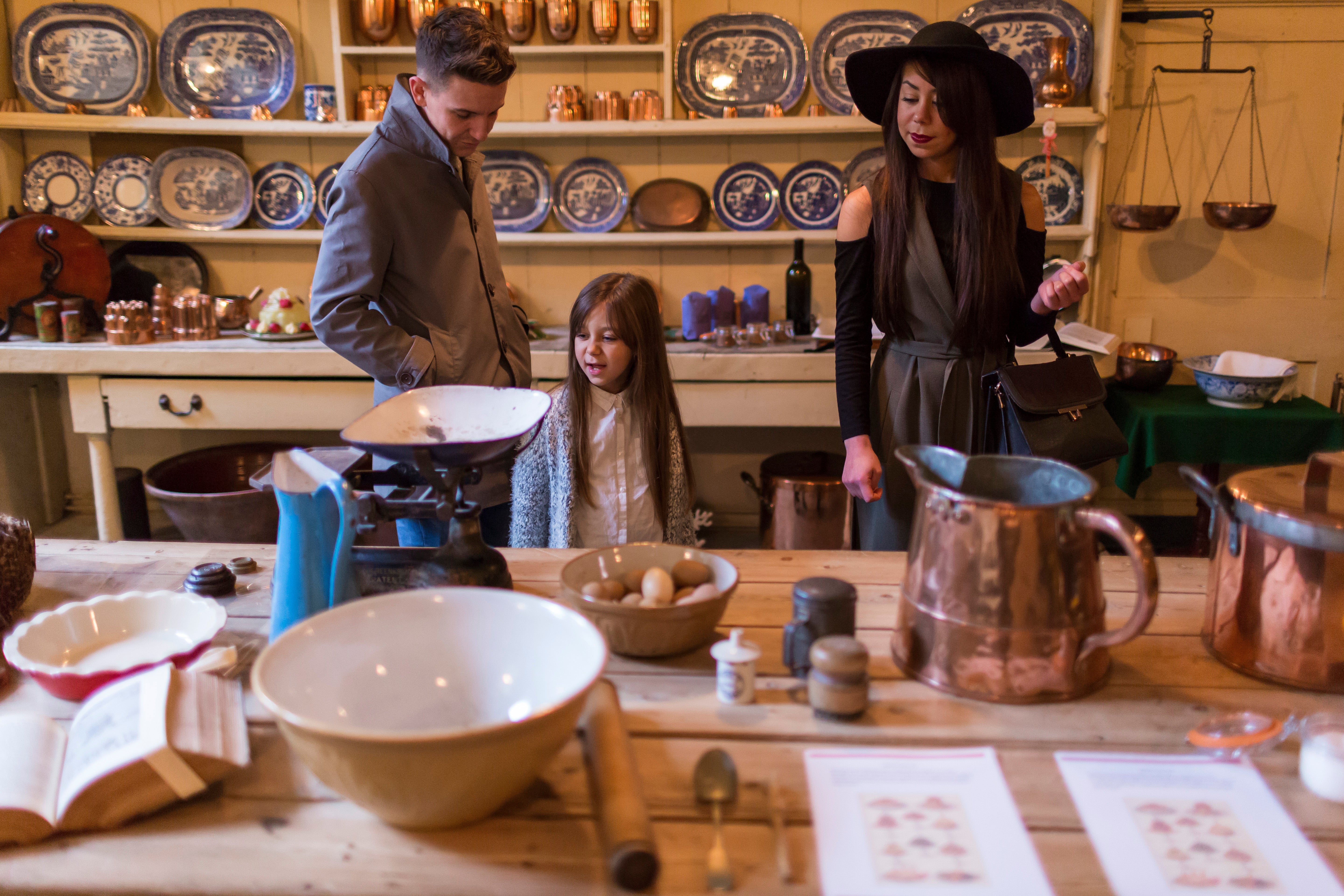 Three visitors stand around a table in a historic kitchen display, examining traditional cooking equipment. The room contains copper pots, ceramic mixing bowls, weighing scales, eggs and kitchen utensils on a wooden worktable, with shelves of blue-and-white plates and copper cookware lining the walls behind them. Warm indoor lighting highlights the period furnishings and collection of household objects.