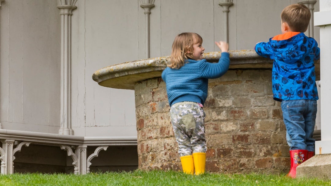 Two children in rainwear standing at brick and stone circular table nside the white painted Gothic pavilion at Uppark, Sussex