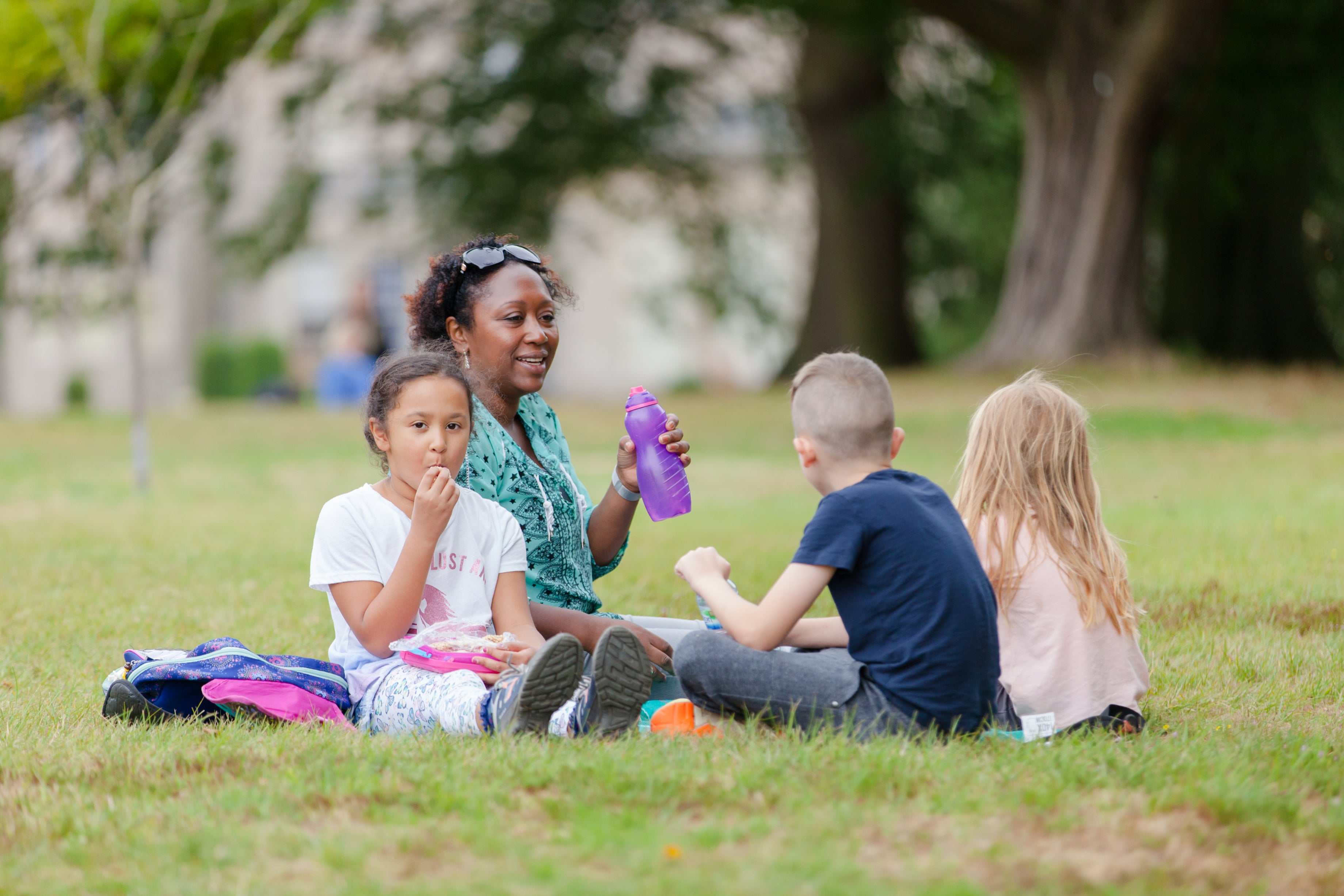 Four people sit together on a grassy lawn in a park, having a picnic. One person holds a purple drinks bottle while a child in the foreground eats from a lunchbox. Two other children sit nearby with their backs partly to the camera. Trees and buildings are softly blurred in the background, and bags and picnic items are placed on the grass.