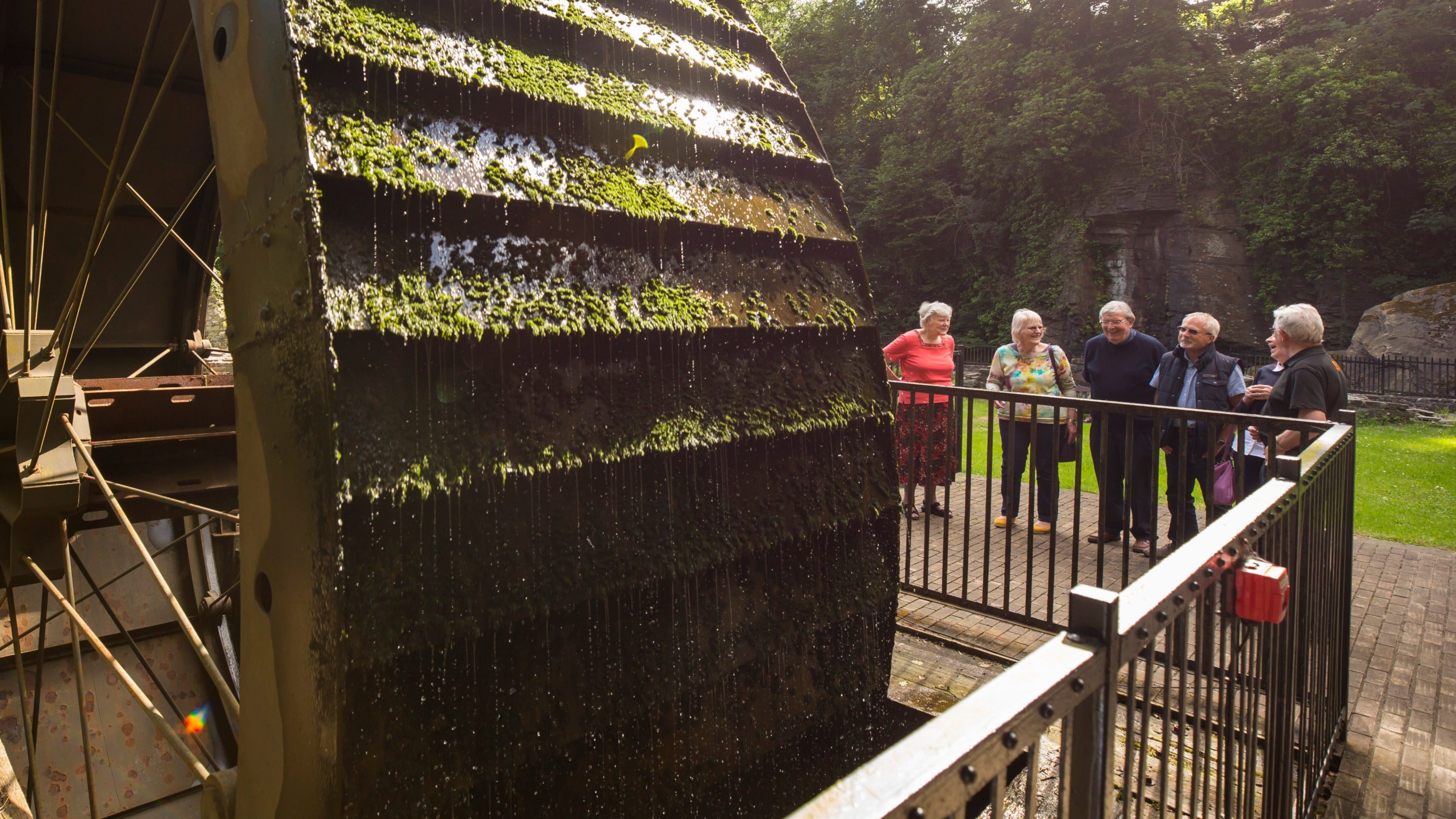 Grŵp o bum oedolyn yn sefyll a gwenu wrth edrych i fyny at olwyn ddŵr fawr, sydd wedi’i gorchuddio ag algâu, yng Ngwaith Tun a Rhaeadr Aberdulais, Cymru.