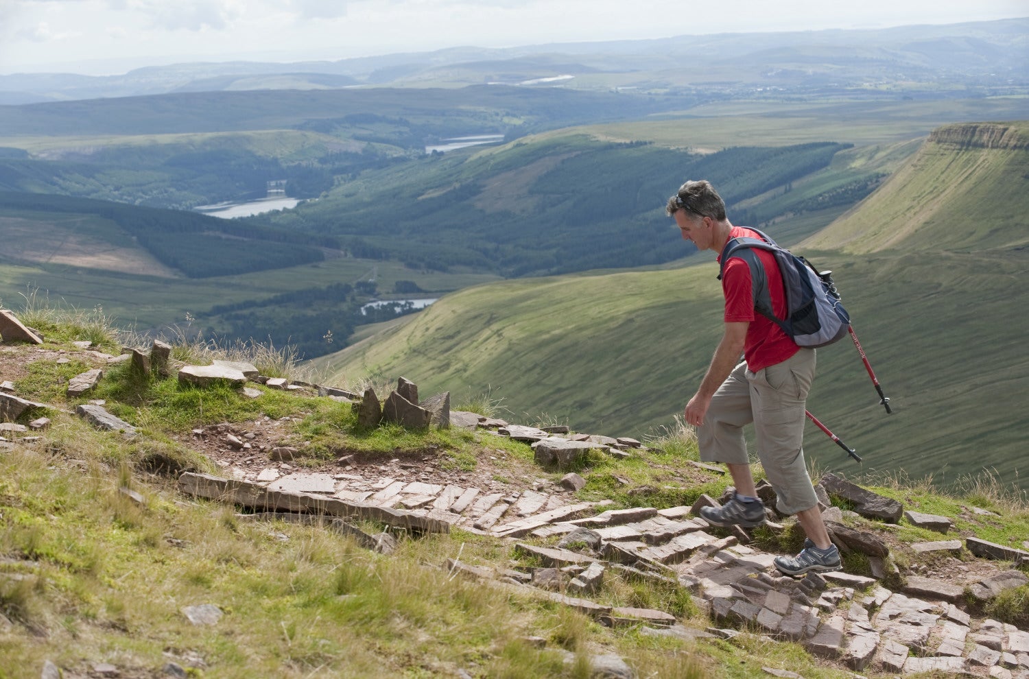 A man walks the Brecon Beacons mountain range, the beautiful scenery pans out in the background as he walks upwards.
