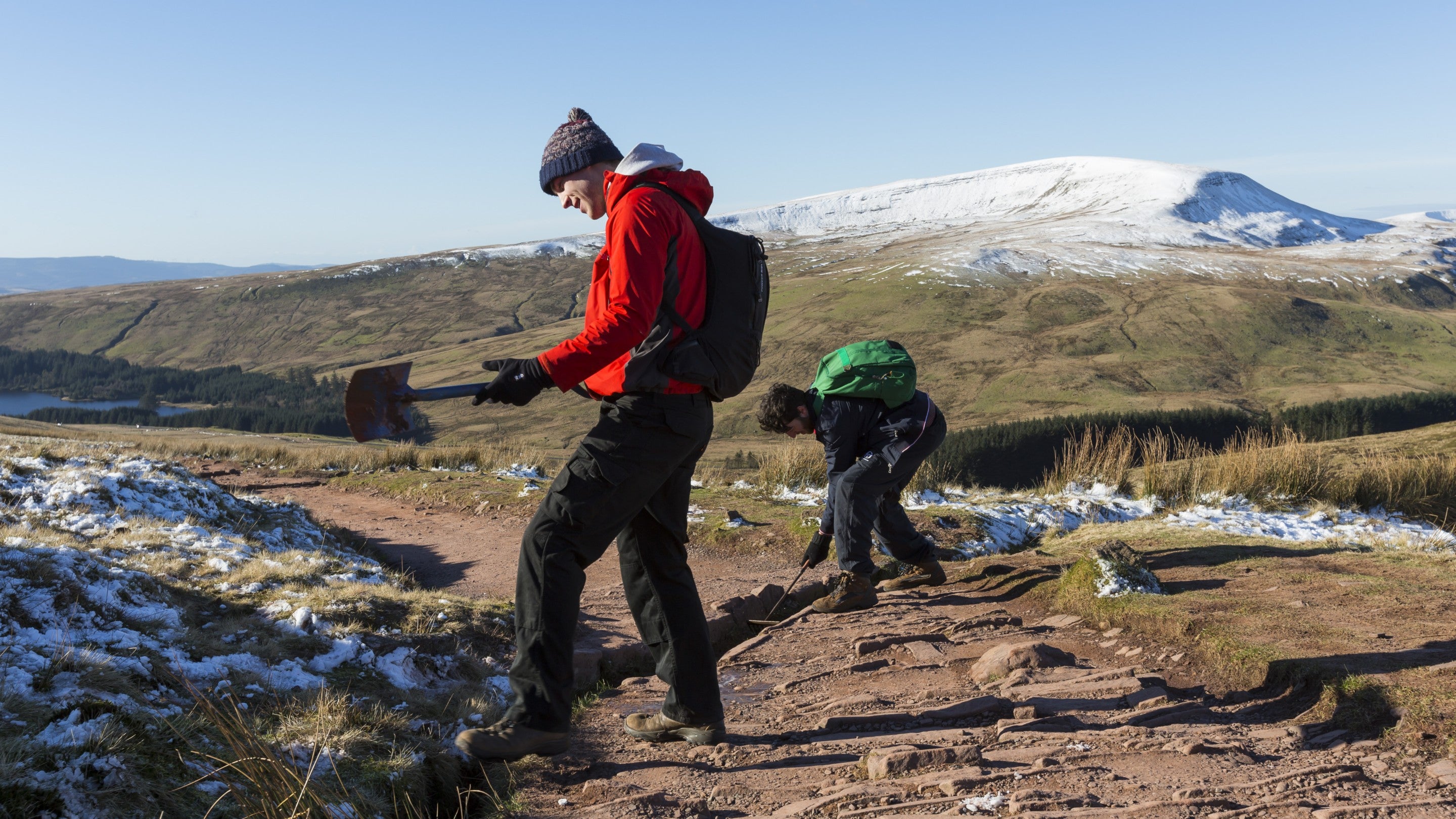 Dau ŵr ifanc yn trwsio cwlfer draenio ar lwybr troed ym Mannau Brycheiniog, gyda gorchudd denau o eira ar y mynyddoedd, ar ddiwrnod braf o aeaf.