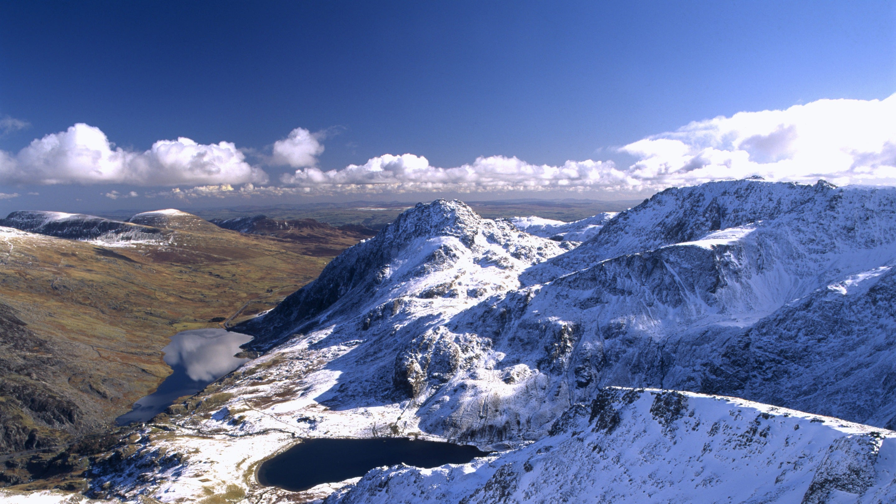 Golygfa o’r uchelderau i lawr o’r Garn ar draws tirwedd mynyddig, dan eira gyda Llyn Ogwen i’w weld yn y dyffryn isod yn Eryri, Cymru.