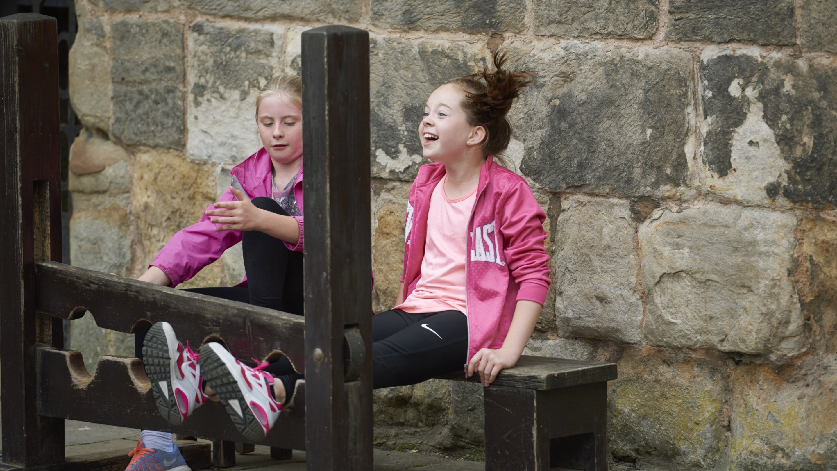 Two children pretending to be stuck in the stocks in the courtyard at Chirk Castle; one with their feet through the foot holes and another child watching.
