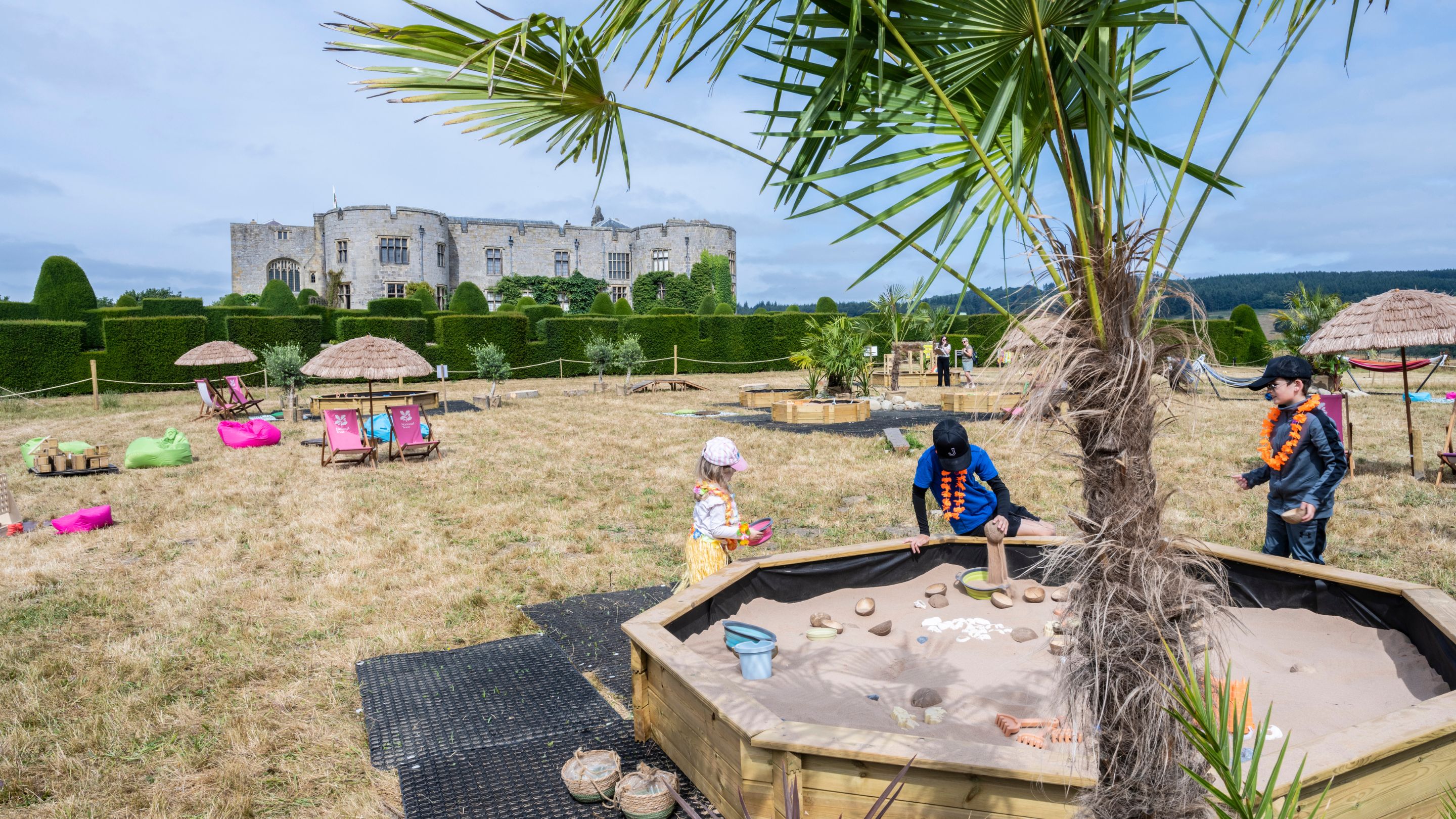 Children play in a large sandpit beneath palm trees at Play in Paradise, with deckchairs, parasols and Chirk Castle in the background.