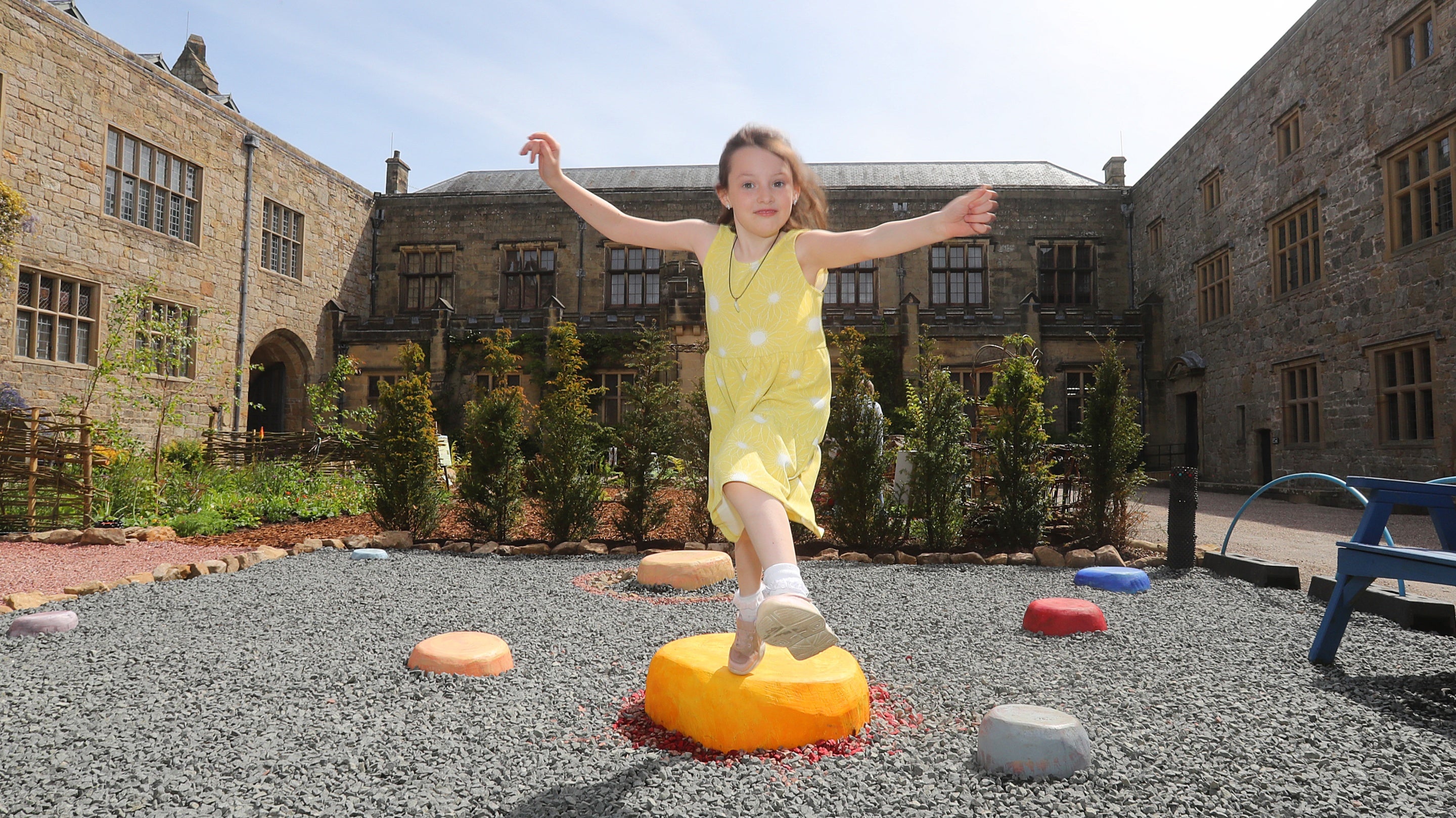 A young girl in a yellow polka dot dress smiles as she jumps across planet 'stepping stones' in the Time-Travel Gardens in the ventral courtyard at Chirk Castle.