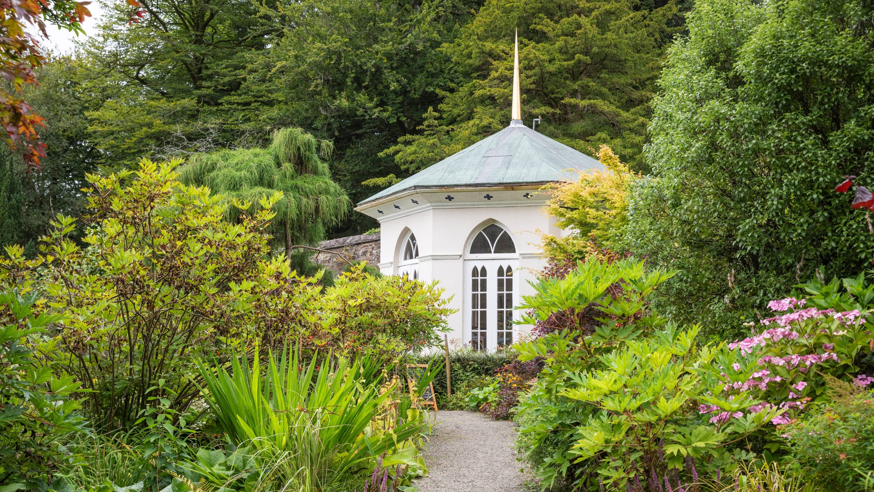 The Colby Gazebo, a small, cream coloured building with tall windows and a pointed roof sites at the end of a garden path which is lined with flowers, plants and trees.