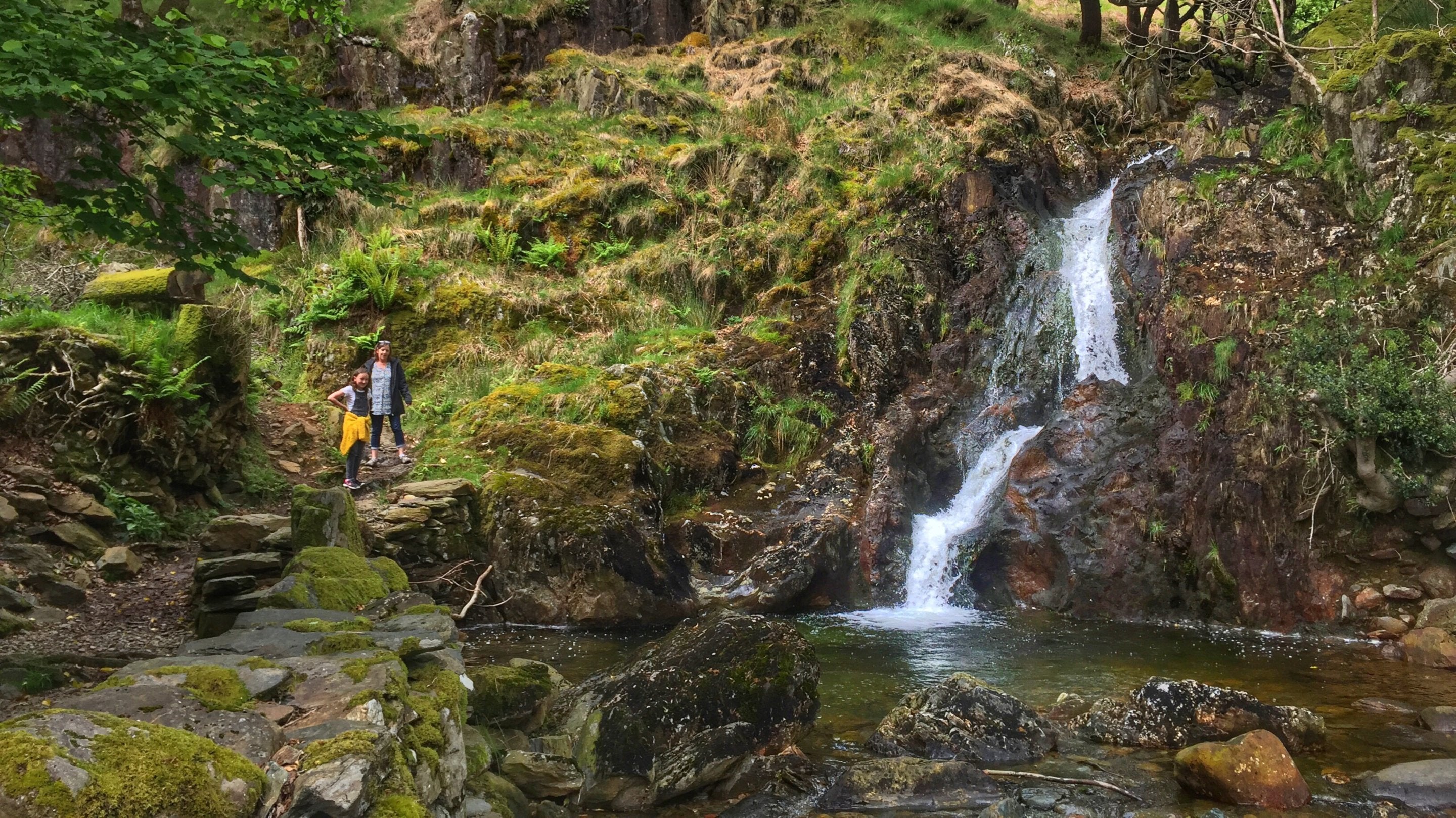 Two people are walking down an uneven grassy slope beside a small waterfall near Dinas Emrys, Beddgelert, Snowdonia, North Wales.