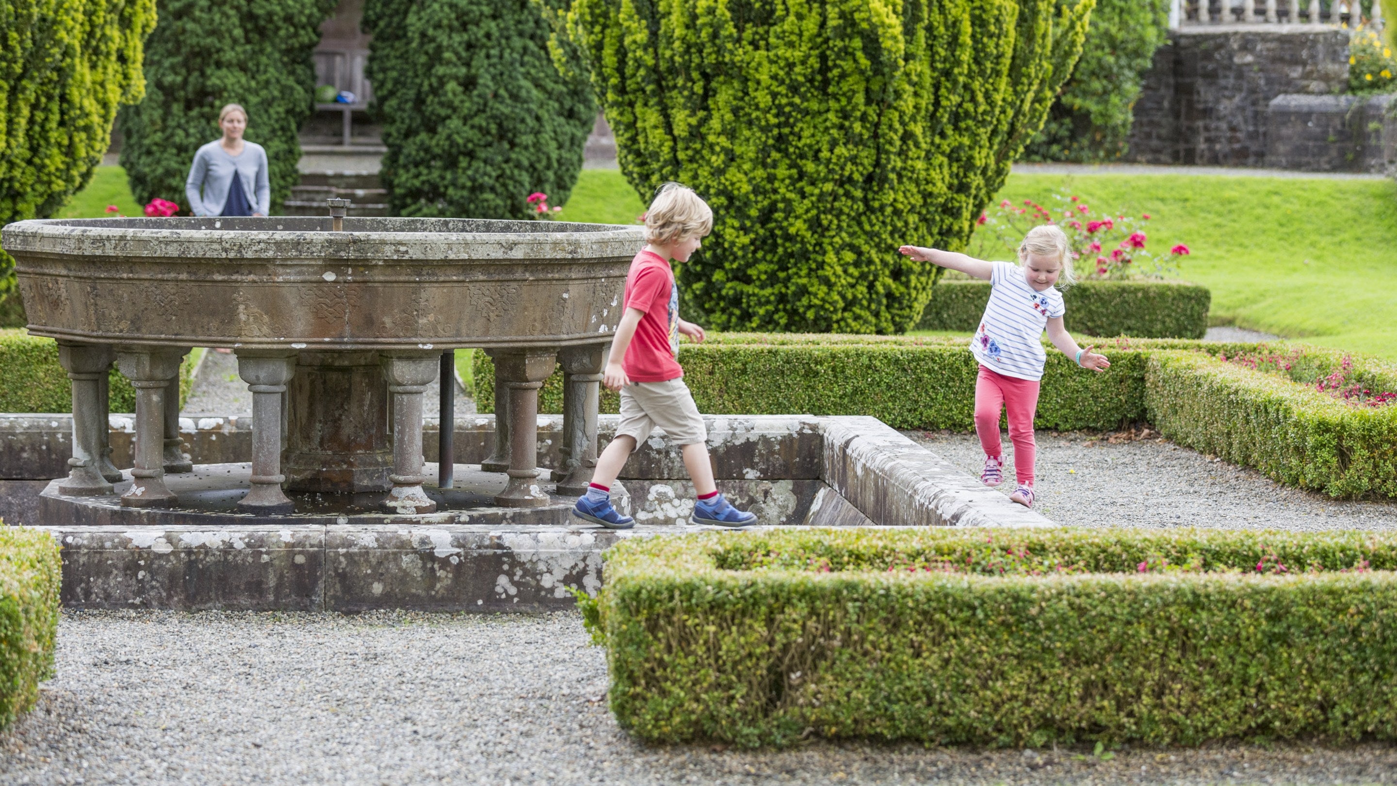Children balance on a low wall around a fountain in the garden at Dinefwr in summer