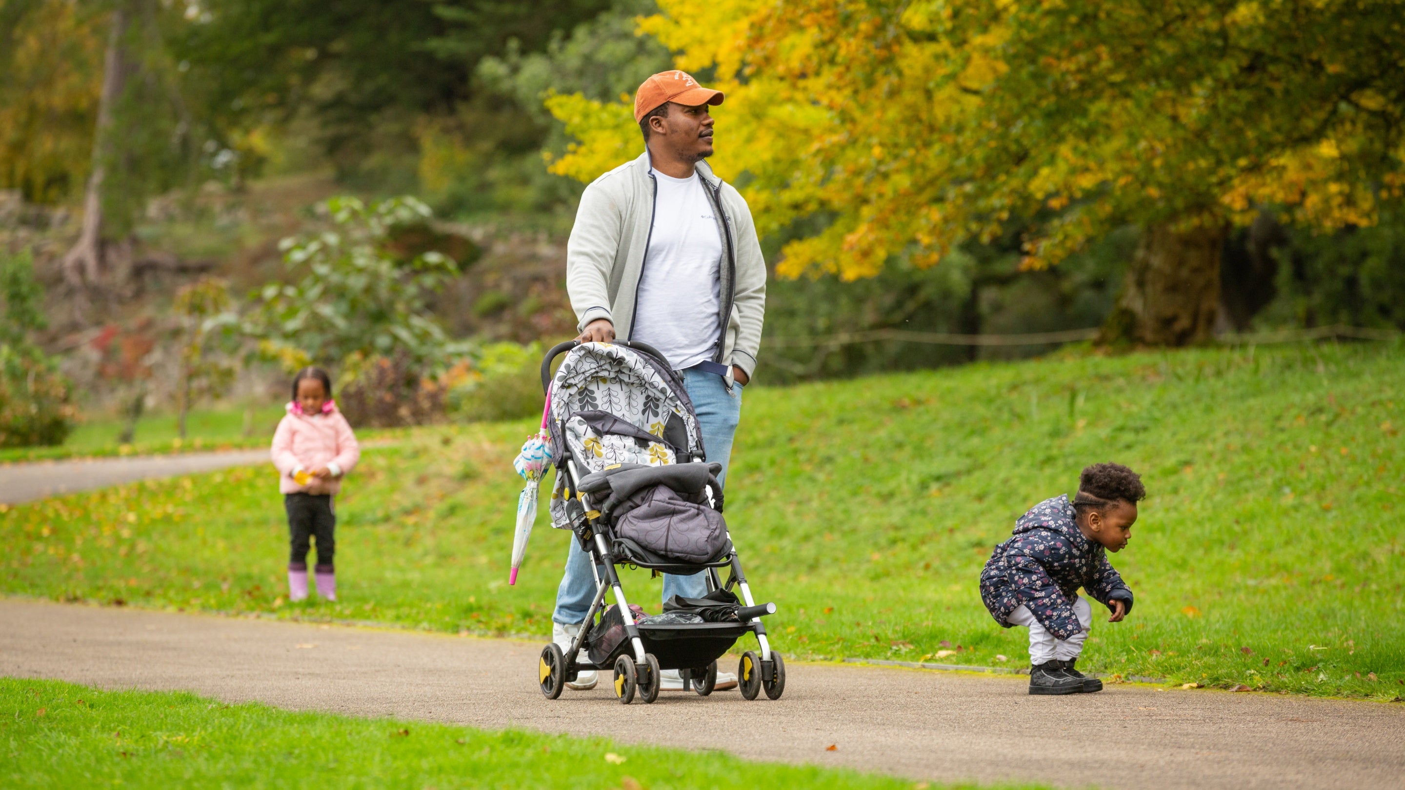 A man pushing a pushchair along a path through lawned gardens with two toddlers – one walking behind and the other in front of him