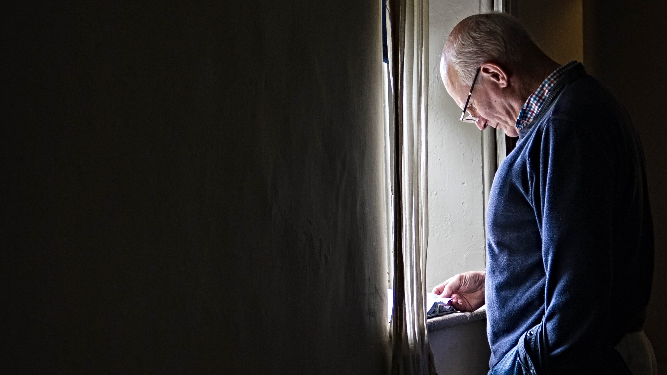 Dyn yn gwisgo sbectol yn sefyll yn darllen wrth ffenestr olau, gyda’r ystafell o’i gwmpas yn dywyllach.