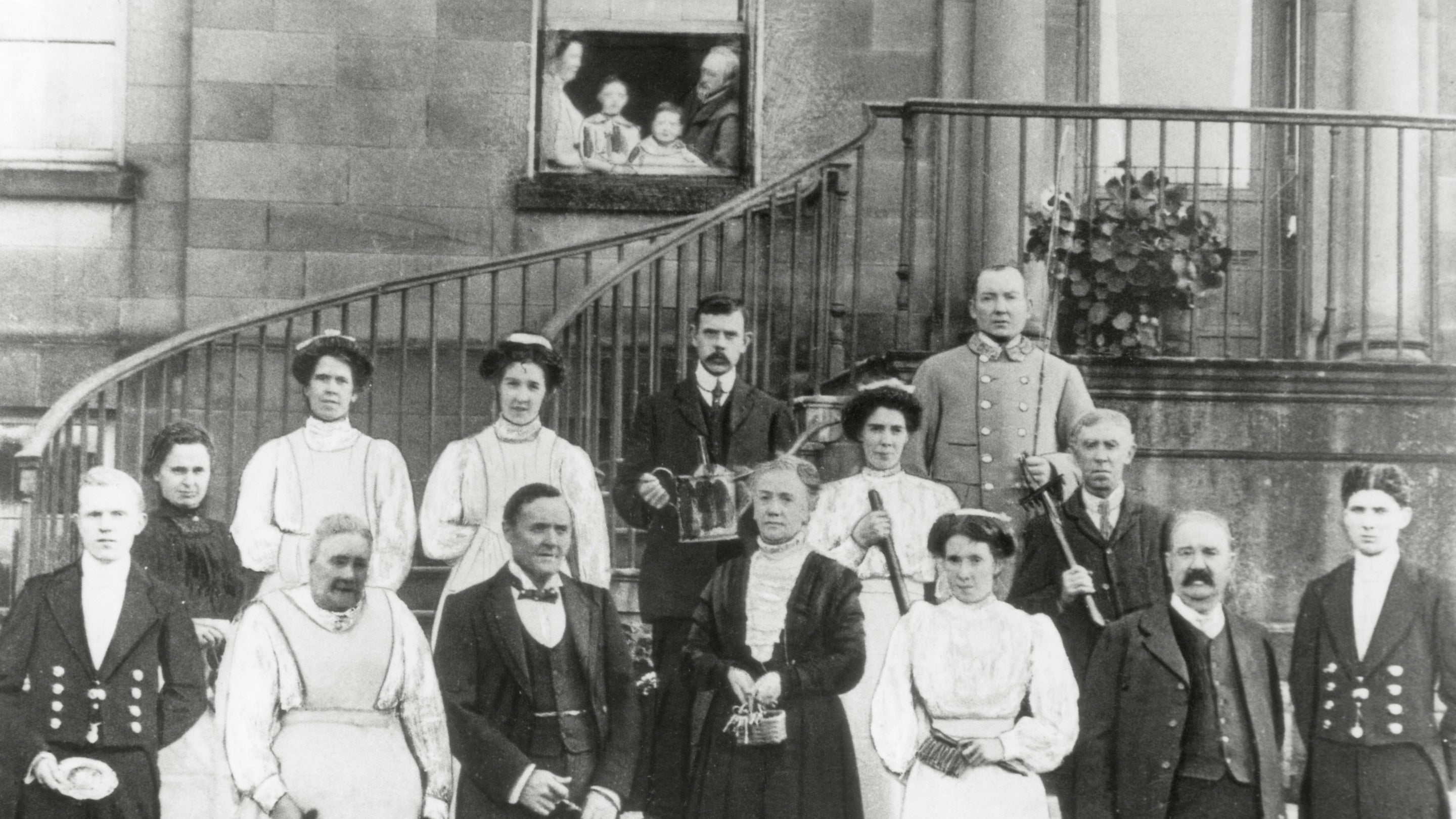 Black and white photo of servants in uniform, standing on the front steps of a grand house. A family of parents and children look on from a first-floor window.