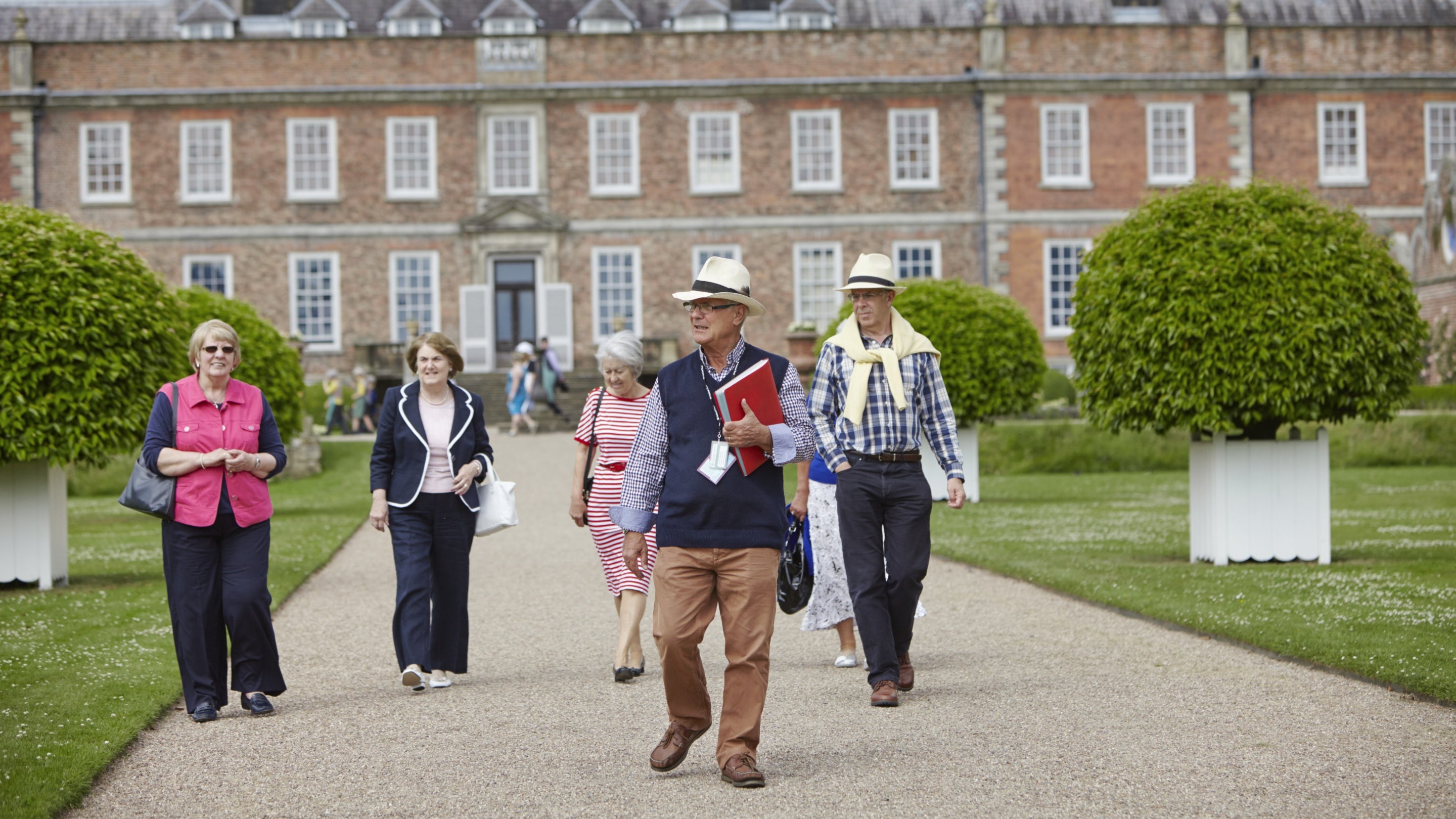 A group of visitors tour a garden in front of a red-brick house, led by a man holding a red folder.