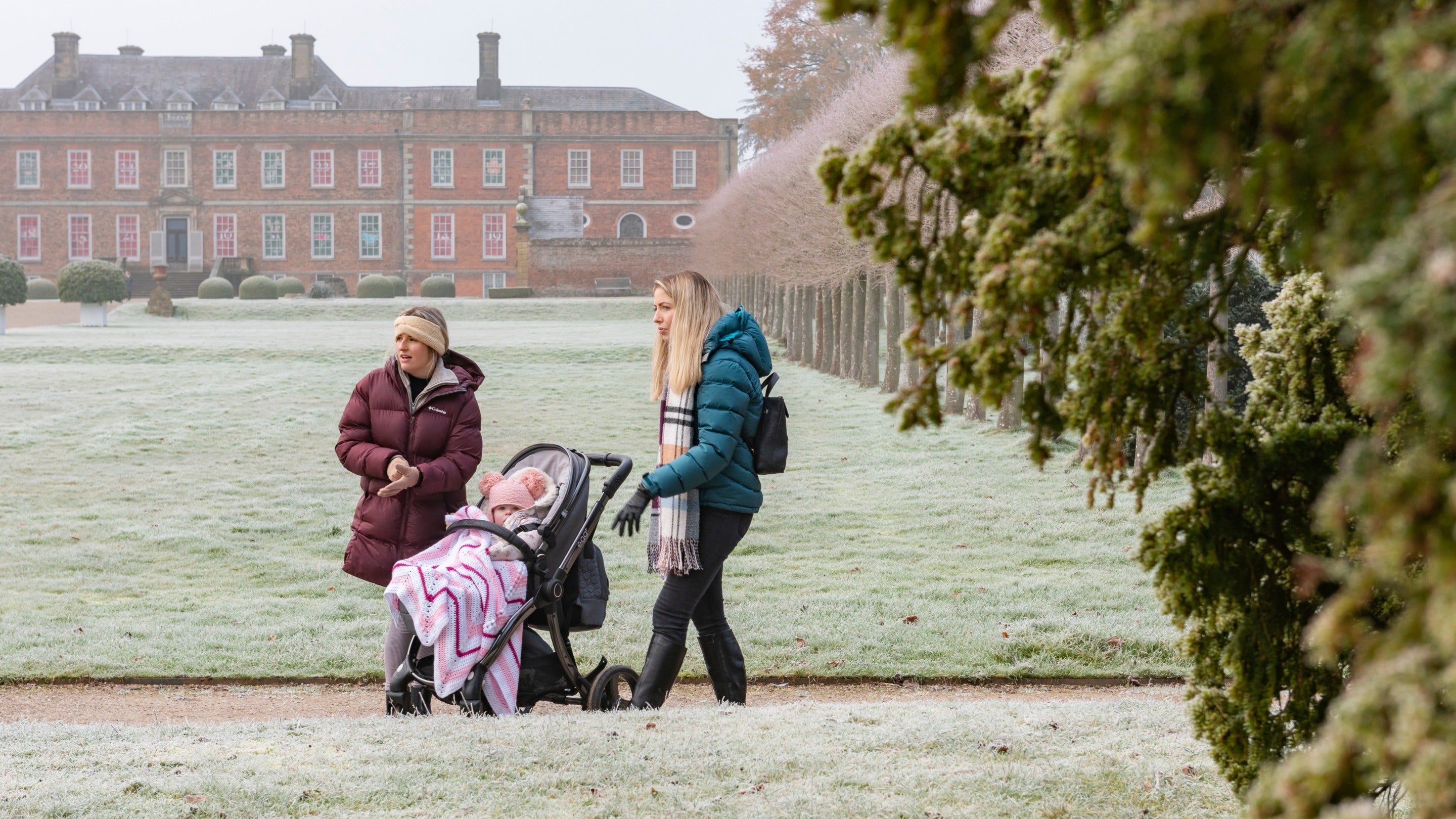 Dwy ddynes gyda phlentyn mewn cadeiriau olwyn yn crwydro gardd Erddig ar ddiwrnod rhewllyd yn y gaeaf, gyda thŷ Erddig yn y cefndir.