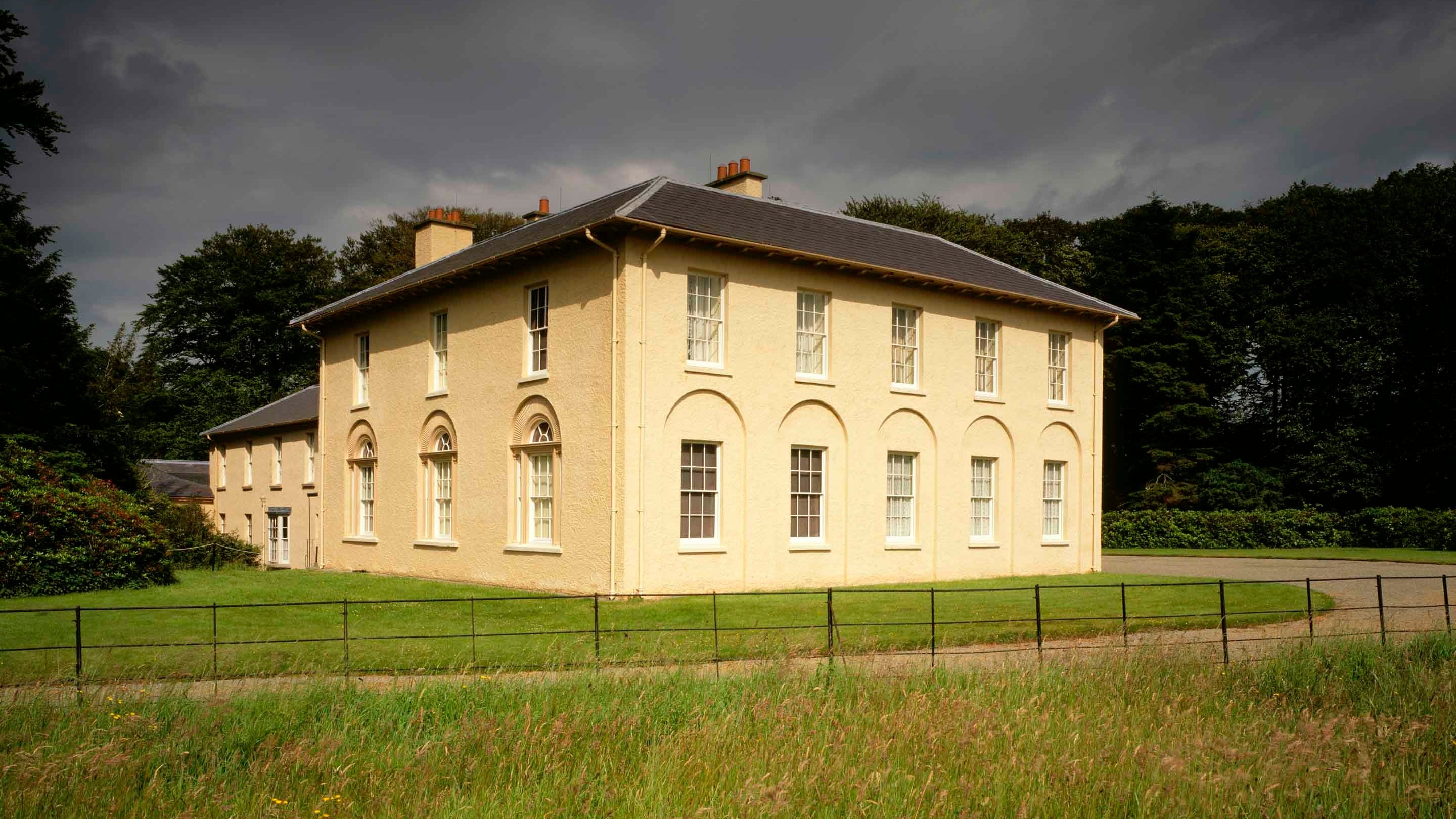 Cornel fila Llanerchaeron, adeilad rendrad hufen gyda tho llechi. Yn y blaendir mae dôl o laswellt hir gyda choed aeddfed y tu ôl i’r tŷ ac awyr lwyd uwchben.