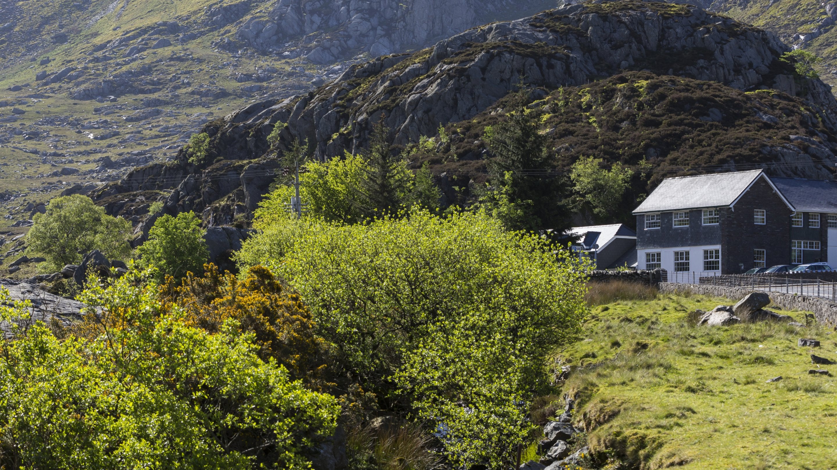 Golygfa o Fwthyn Ogwen, y Carneddau a’r Glyderau, Gwynedd a Chwm Idwal yn y cefndir. Mae’r bwthyn yn adeilad du a gwyn mawr a saif ar lan nant mewn cwm creigiog.