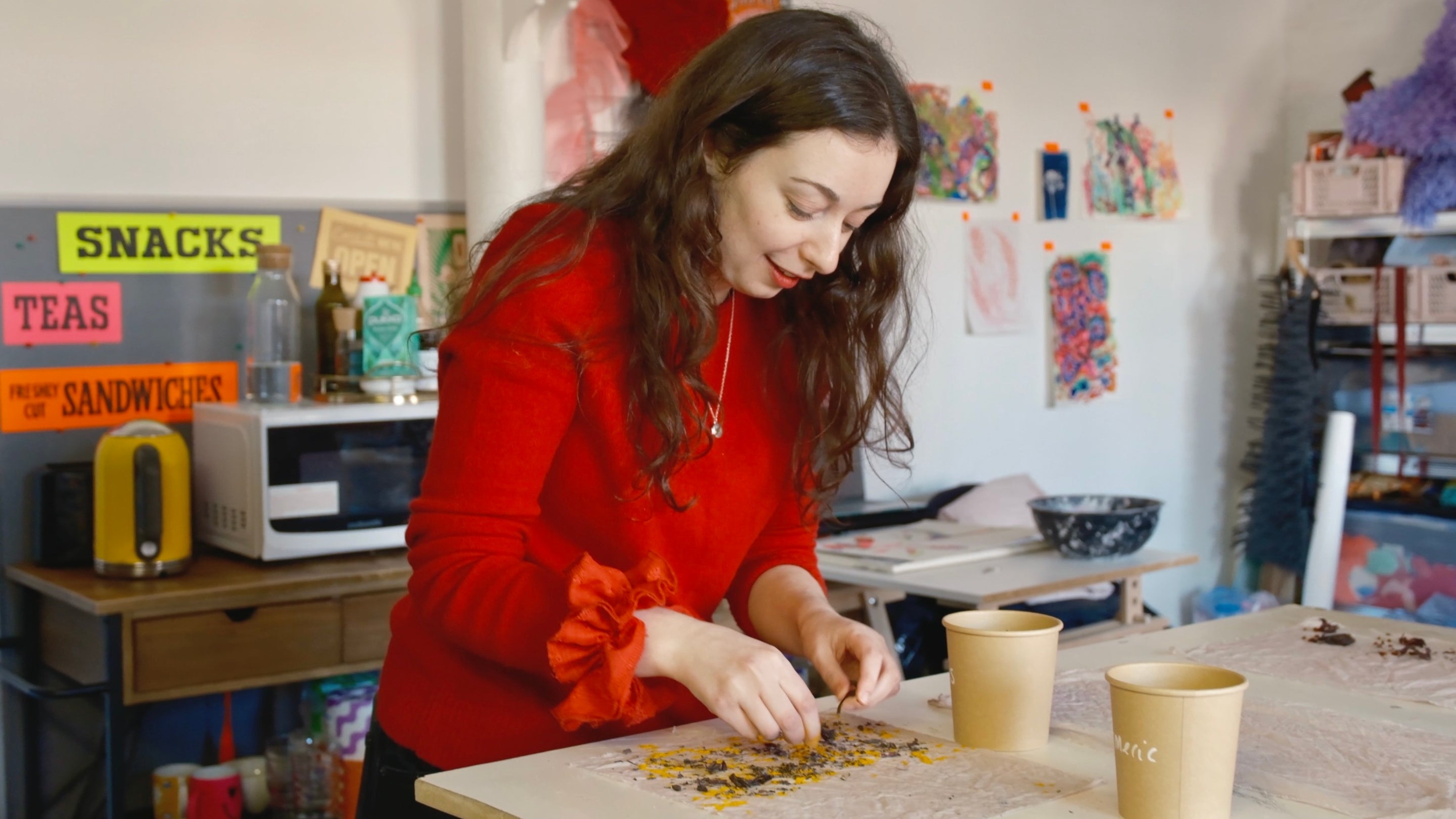 This image shows a woman in a red sweater working on an art project in a creative studio space. She appears to be arranging or pressing dried flowers or leaves onto a flat surface, possibly making handmade paper or a botanical artwork. The environment around her suggests a lively and artistic atmosphere with colourful artwork pinned on the wall.