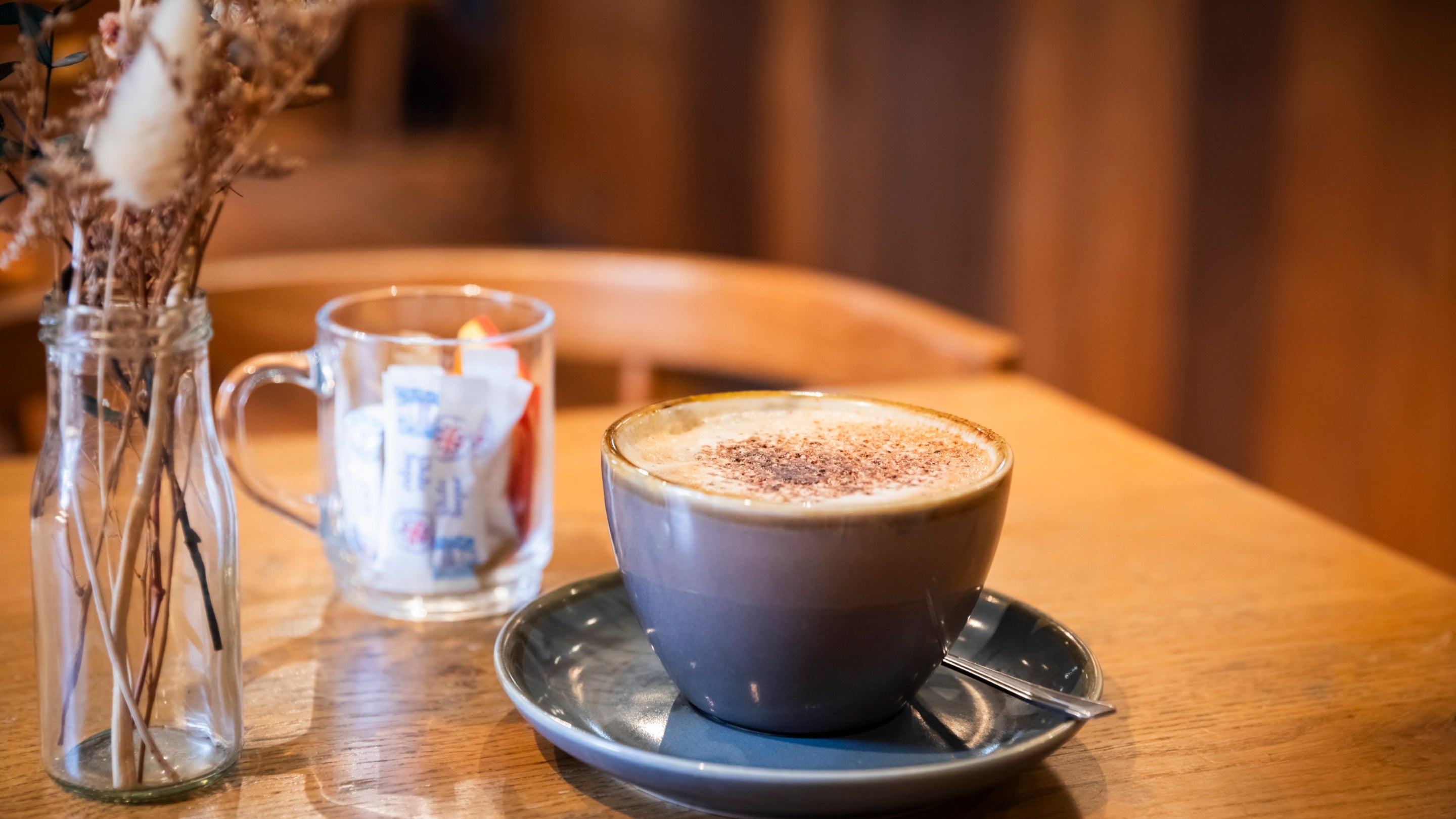A cup of coffee in the forefront on a wooden table with sugar packets in a glass behind