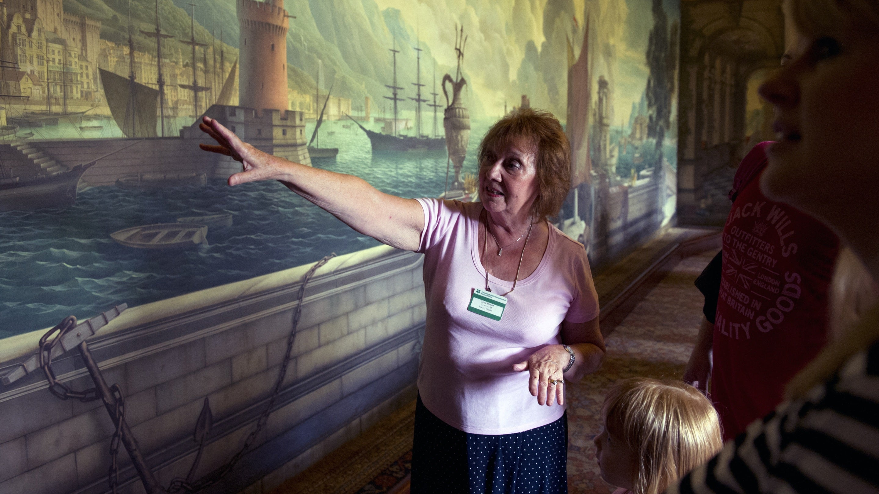 Gwirfoddolwr yn dangos y murlun Rex Whistler i ymwelwyr yn yr Ystafell Fwyta ym Mhlasty a Gerddi Plas Newydd, Ynys Môn, Cymru