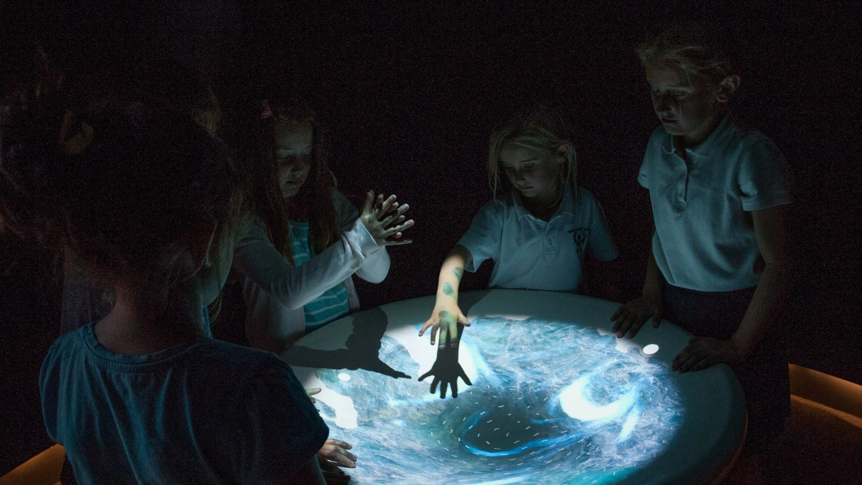 Grŵp o blant yn sefyll o gwmpas arddangosiad crwn mewn ystafell dywyll ym Mhorth y Swnt, Aberdaron, Gwynedd. Mae’r arddangosiad wedi ei oleuo ac mae gwawl las yn cael ei adlewyrchu ar wynebau’r plant.