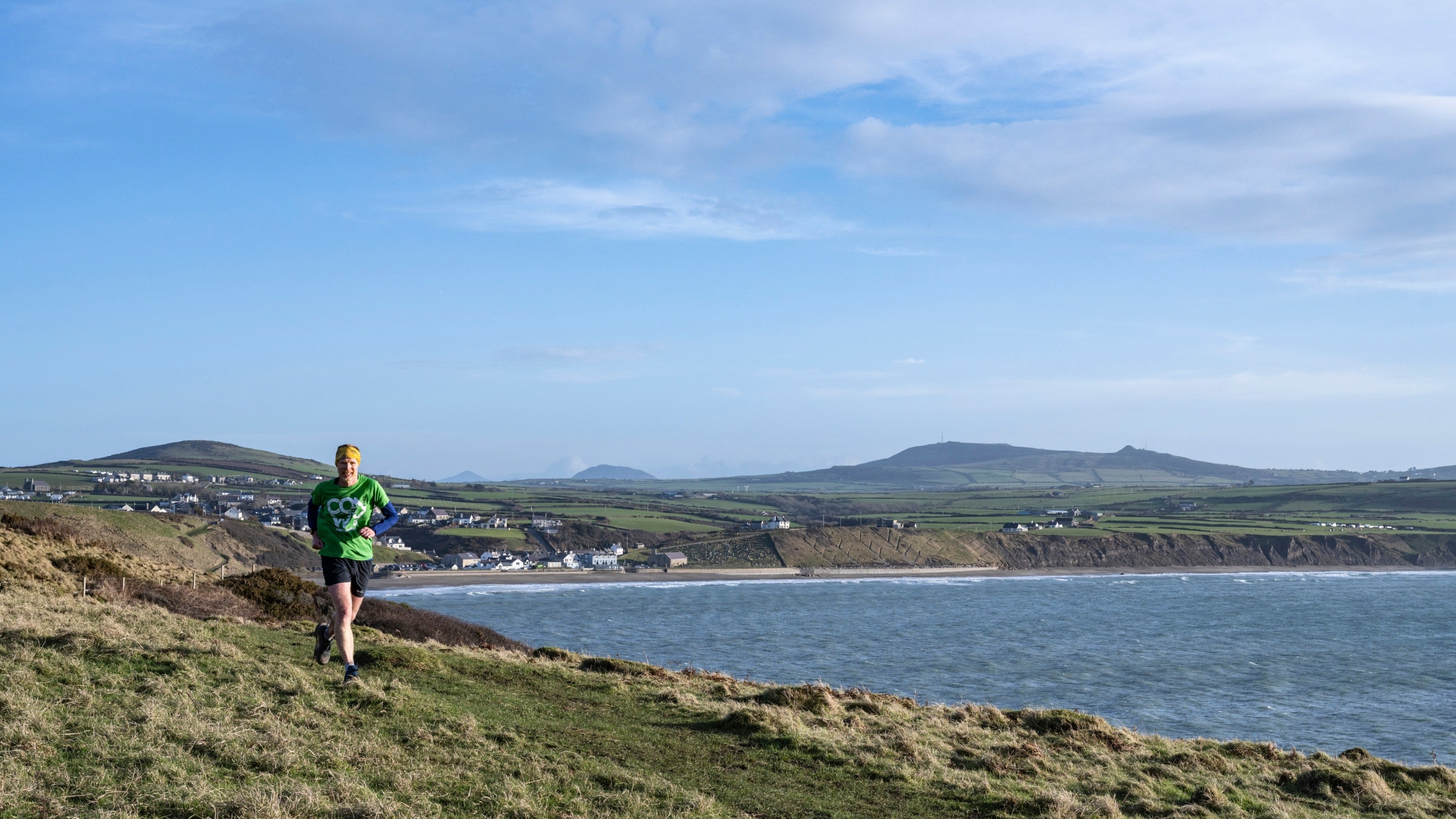 A runner wearing a green top and black shorts with a view down and across Aberdaron during a Trust10 run