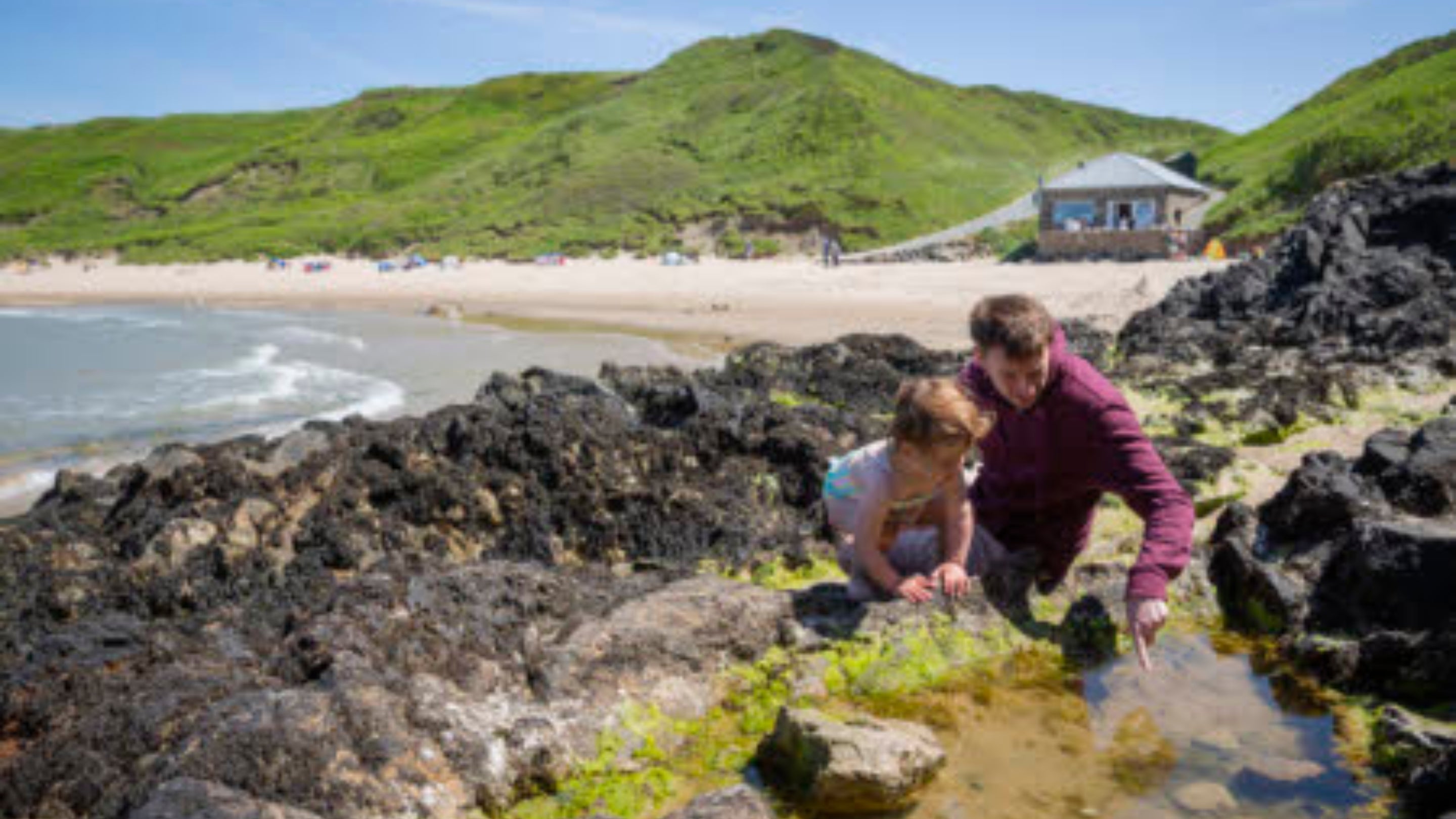 A man and young girl look into a rock pool with a beach and blue skies behind