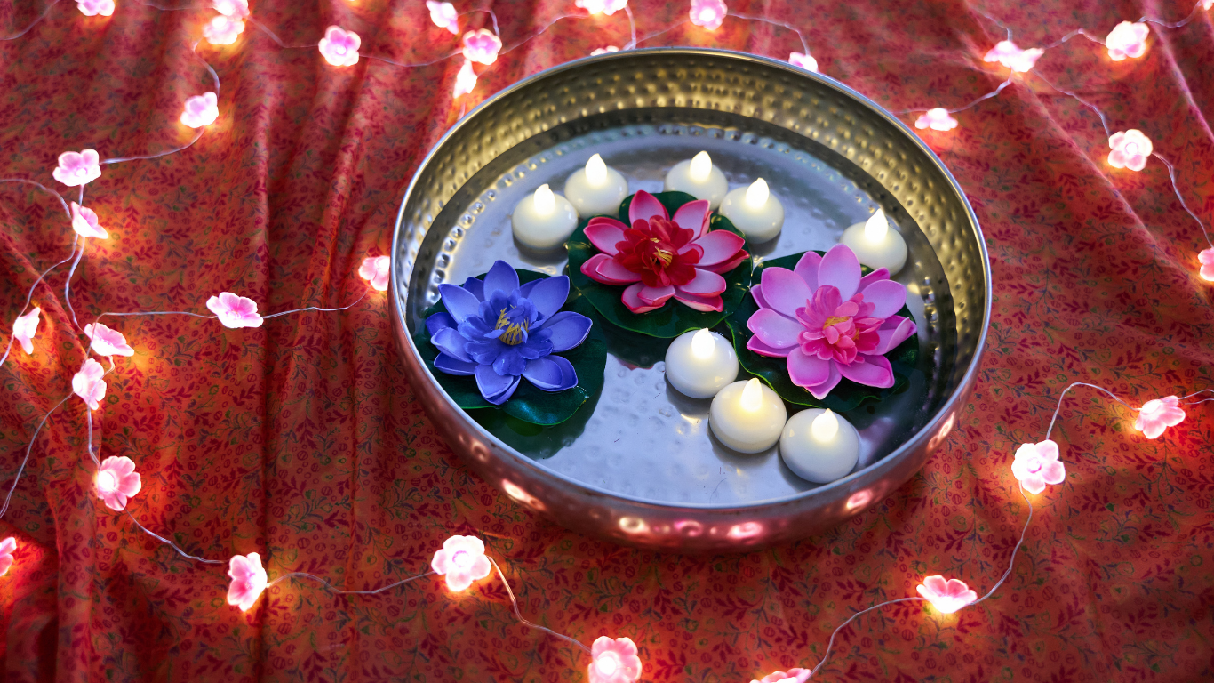 Metal bowl with floating pink and blue water lilies, surrounded by glowing white tea lights and flower-shaped fairy lights on a red patterned cloth.