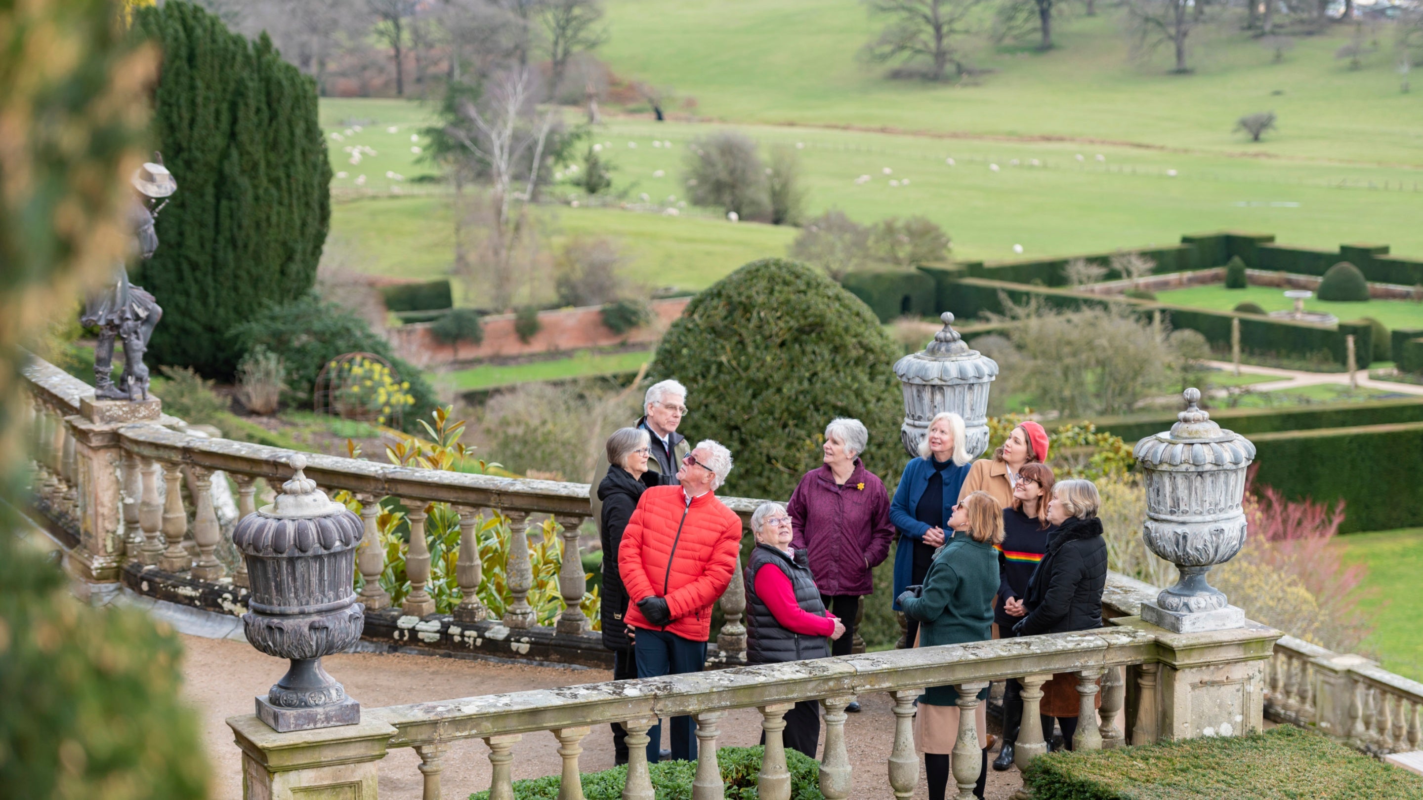 Several people stand on a stone terrace behind ornamental features with green winter landscape in the background