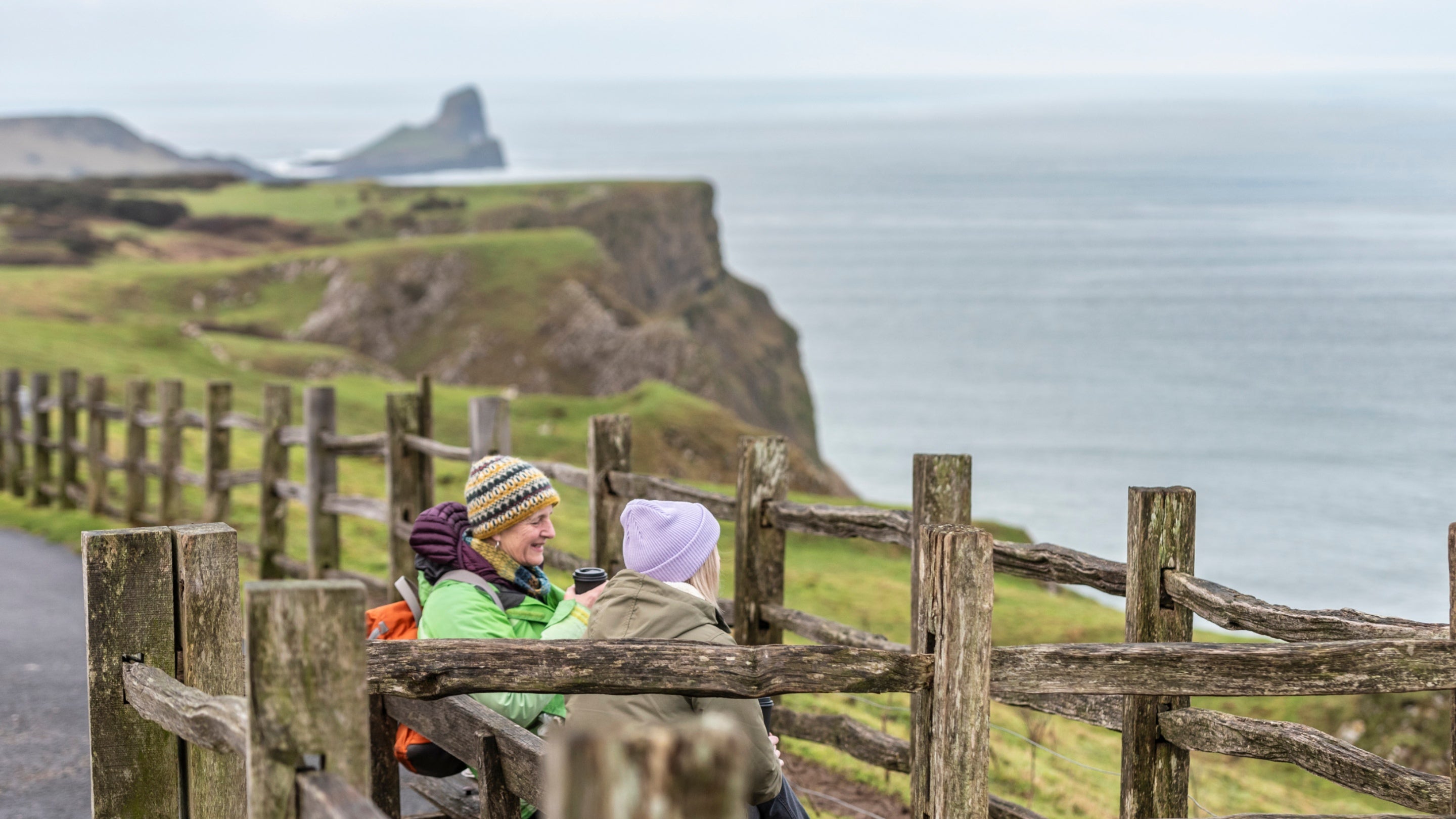 Two people sat on a bench wearing coats and hats. There is a wooden fence between them and they are overlooking the see with the hills in the background