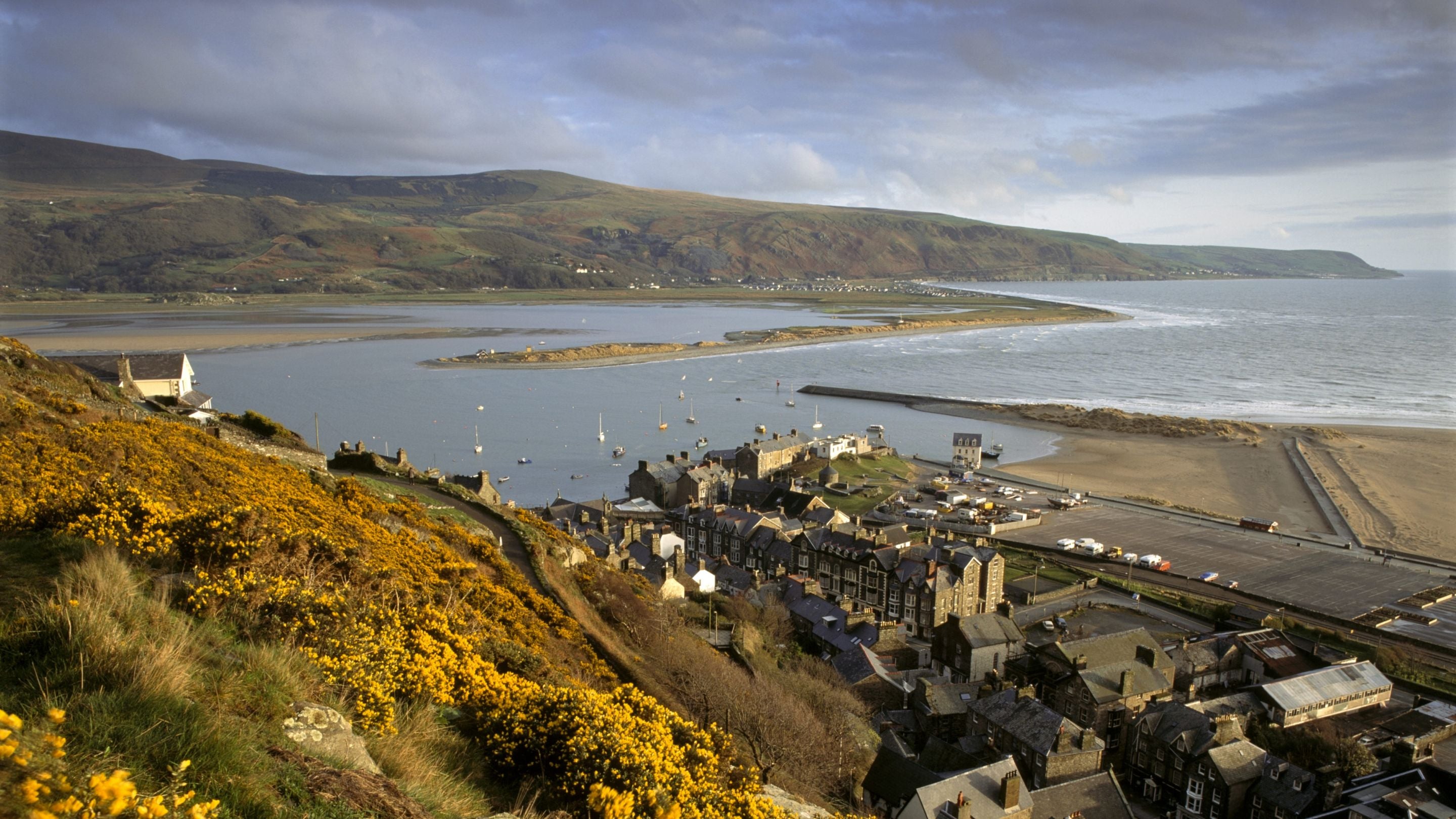 Golygfa i lawr tuag at Abermaw, Gwynedd, o’r clogwyni llawn eithin yn Ninas Oleu. Tu hwnt i doeau’r dref, gwelir y traeth a’r môr tu hwnt iddo.