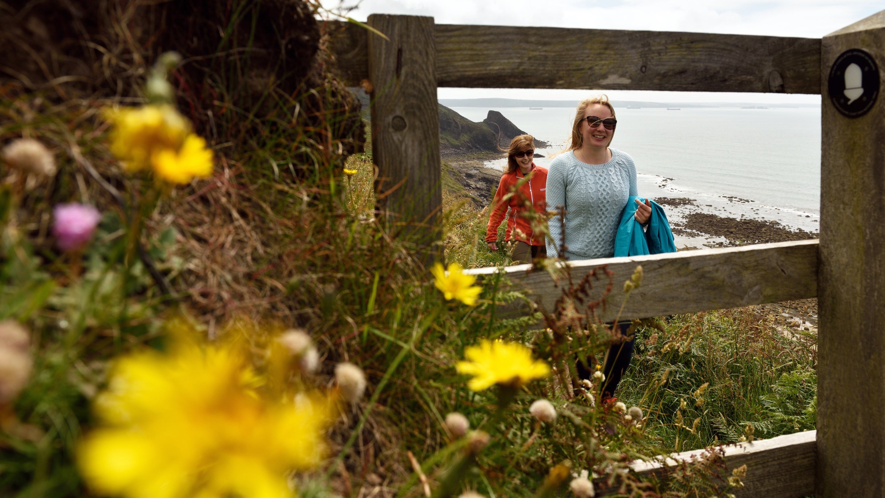 Dwy fenyw’n nesáu at gât ar lwybr yr arfordir yn Stad Southwood, gyda banc o flodau gwyllt yn y blaendir