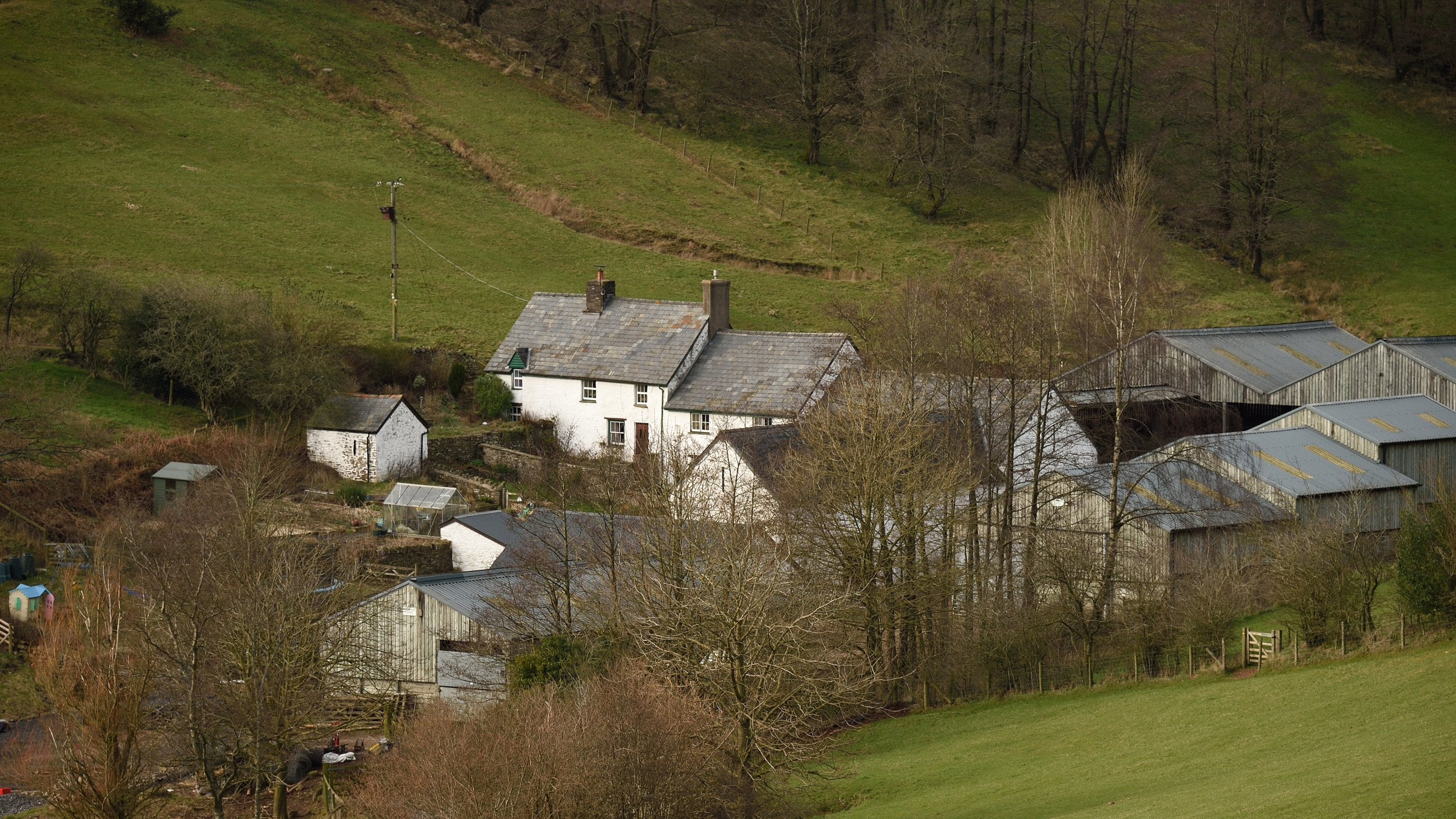 Golygfa o uchder o Fferm Parc Lodge ym Mannau Brycheiniog, Powys, casgliad o sawl adeilad gan gynnwys ffermdy gwyn. Mae’n sefyll mewn pant yn y dirwedd, ac mae nifer o goed heb ddail yn amgylchynu’r eiddo.