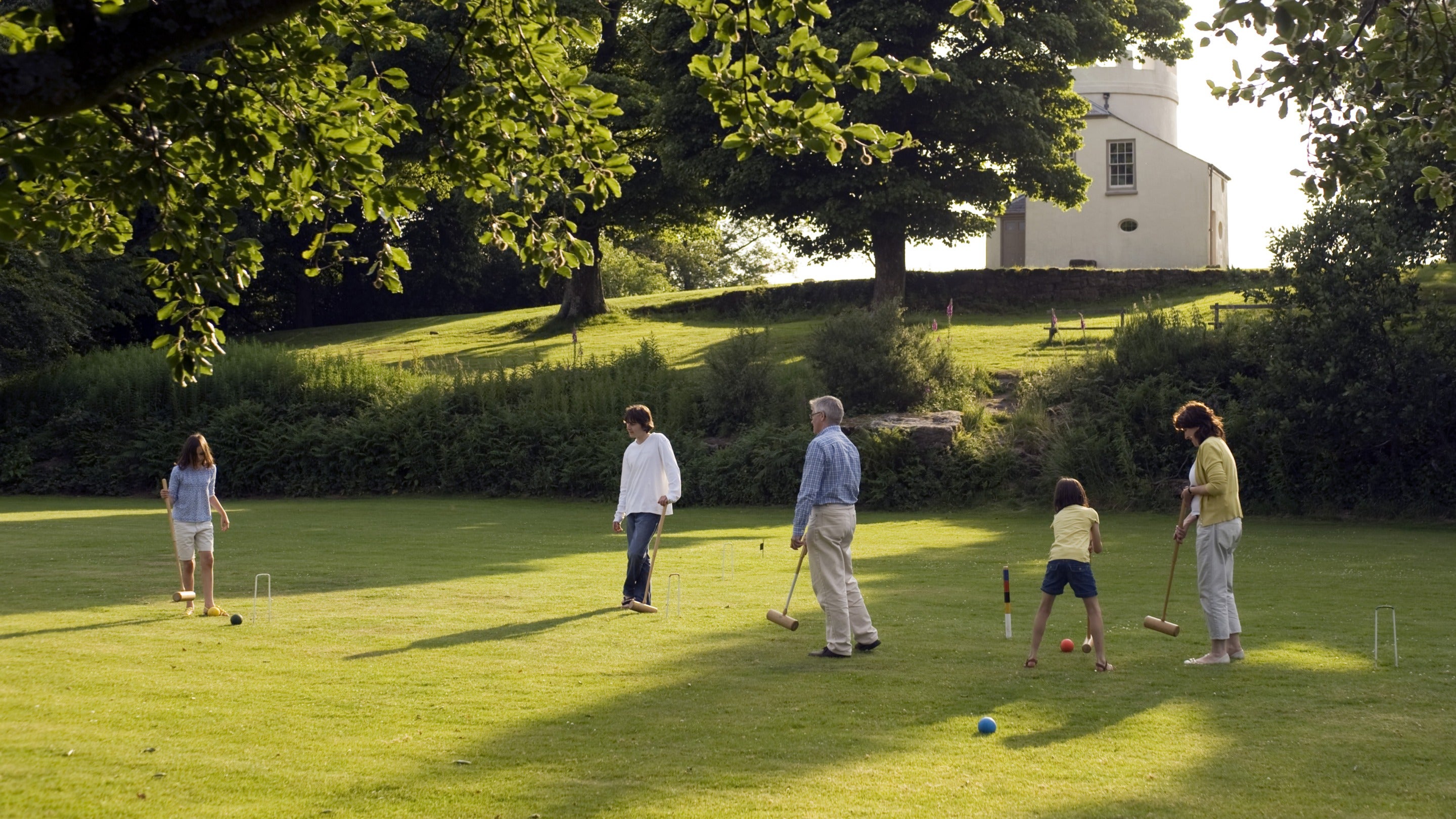 A family of five is playing croquet on a well-kept lawn at The Kymin, Monmouthshire, with the Round House visible through the trees and up a hill in the background.