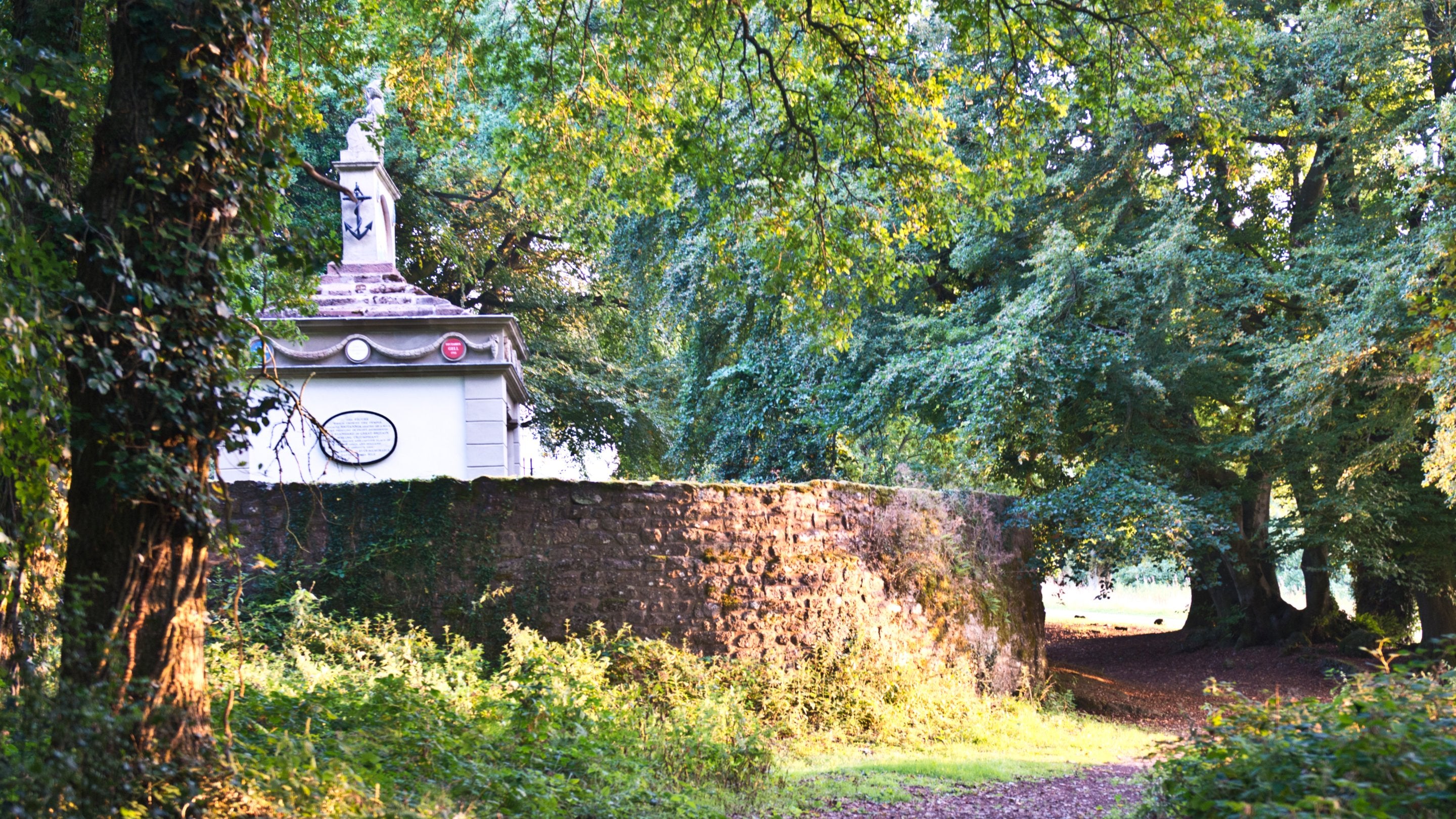 A woodland path with overhanging trees leading towards the Naval Temple at The Kymin, Monmouthshire, which can be partially seen behind a stone wall.