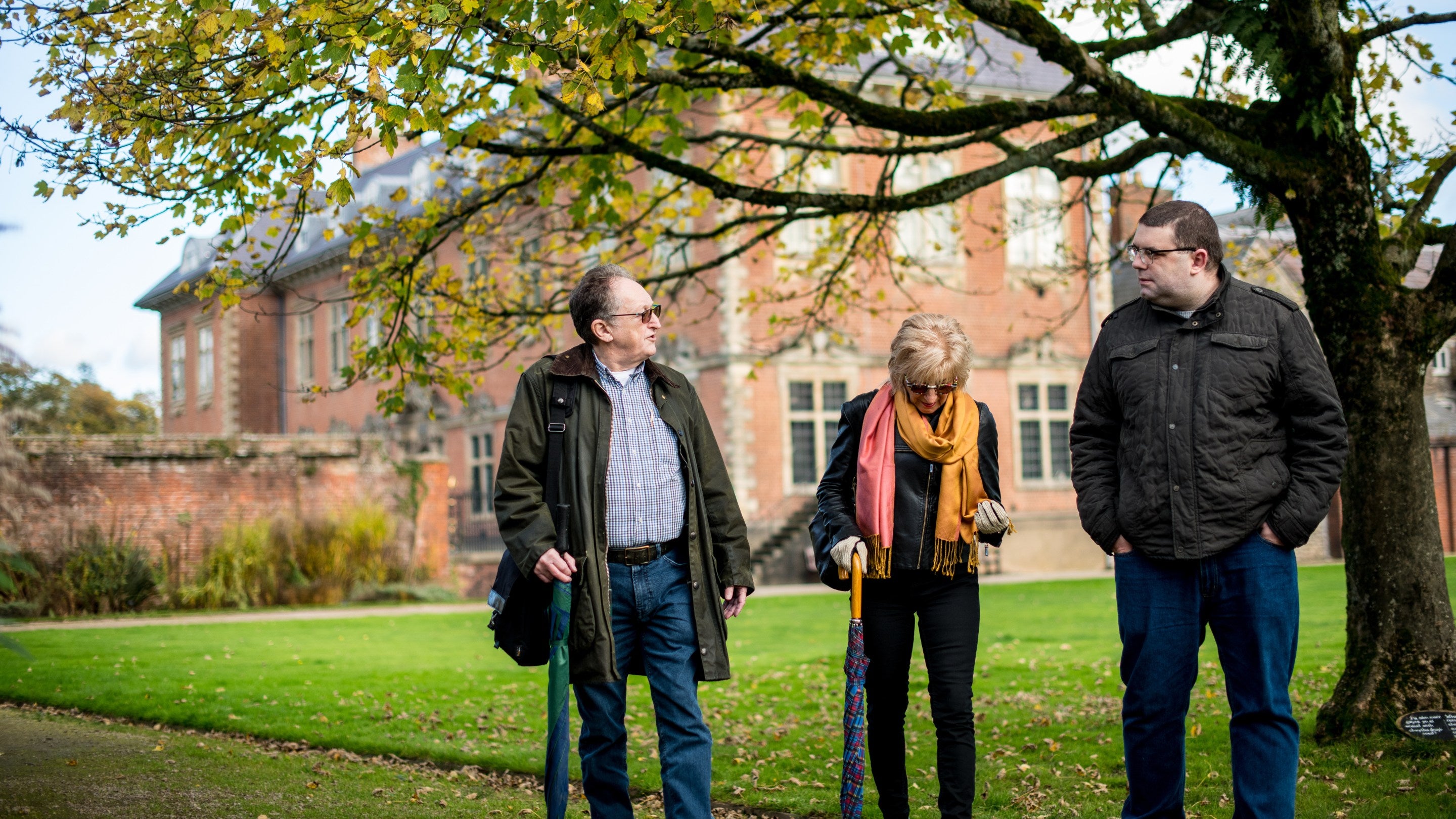Three visitors chat under an acer tree in front of the red-brick mansion house