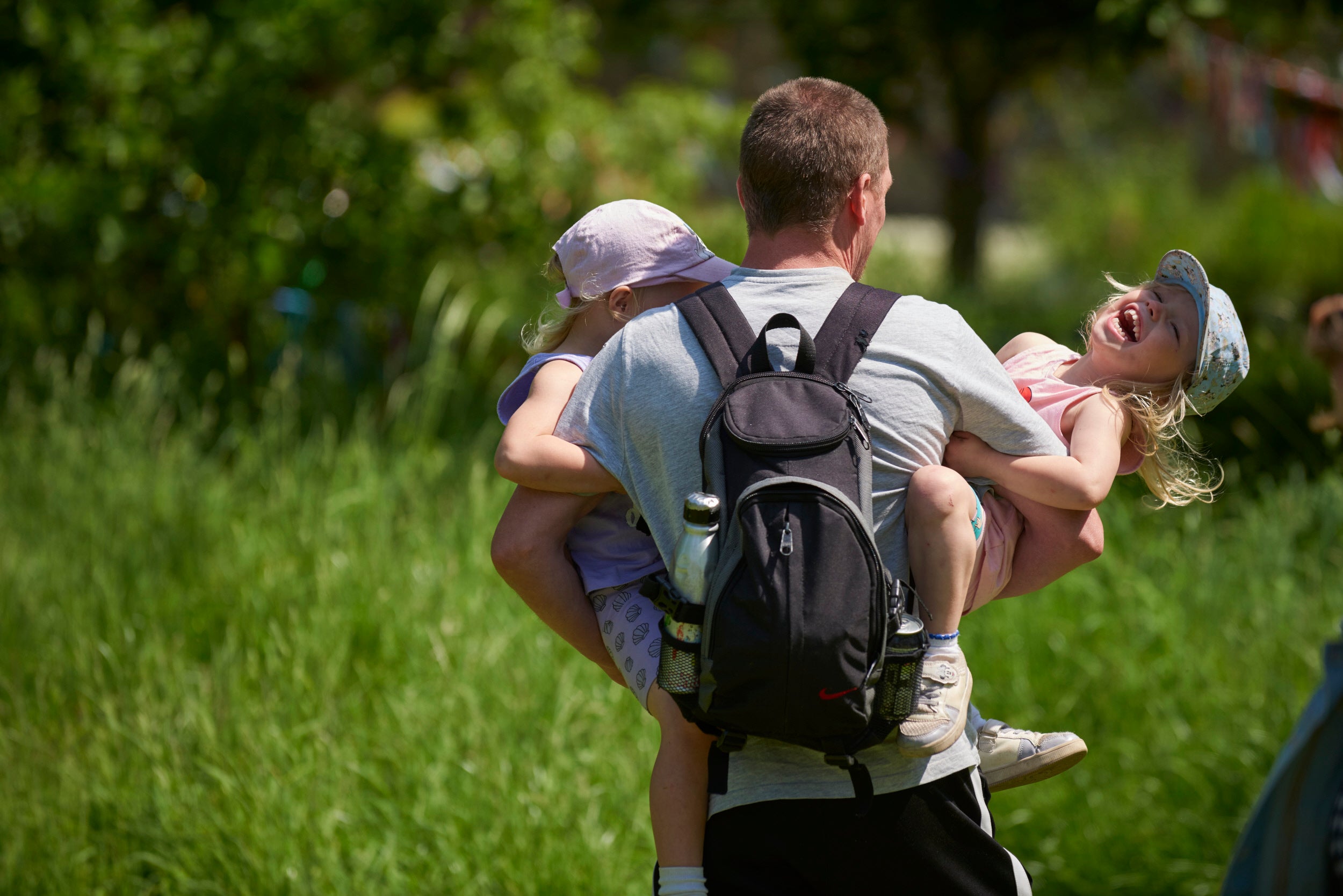 A man facing away from the camera with a backpack on carrying two small children who are both smiling. It is a sunny summer day.