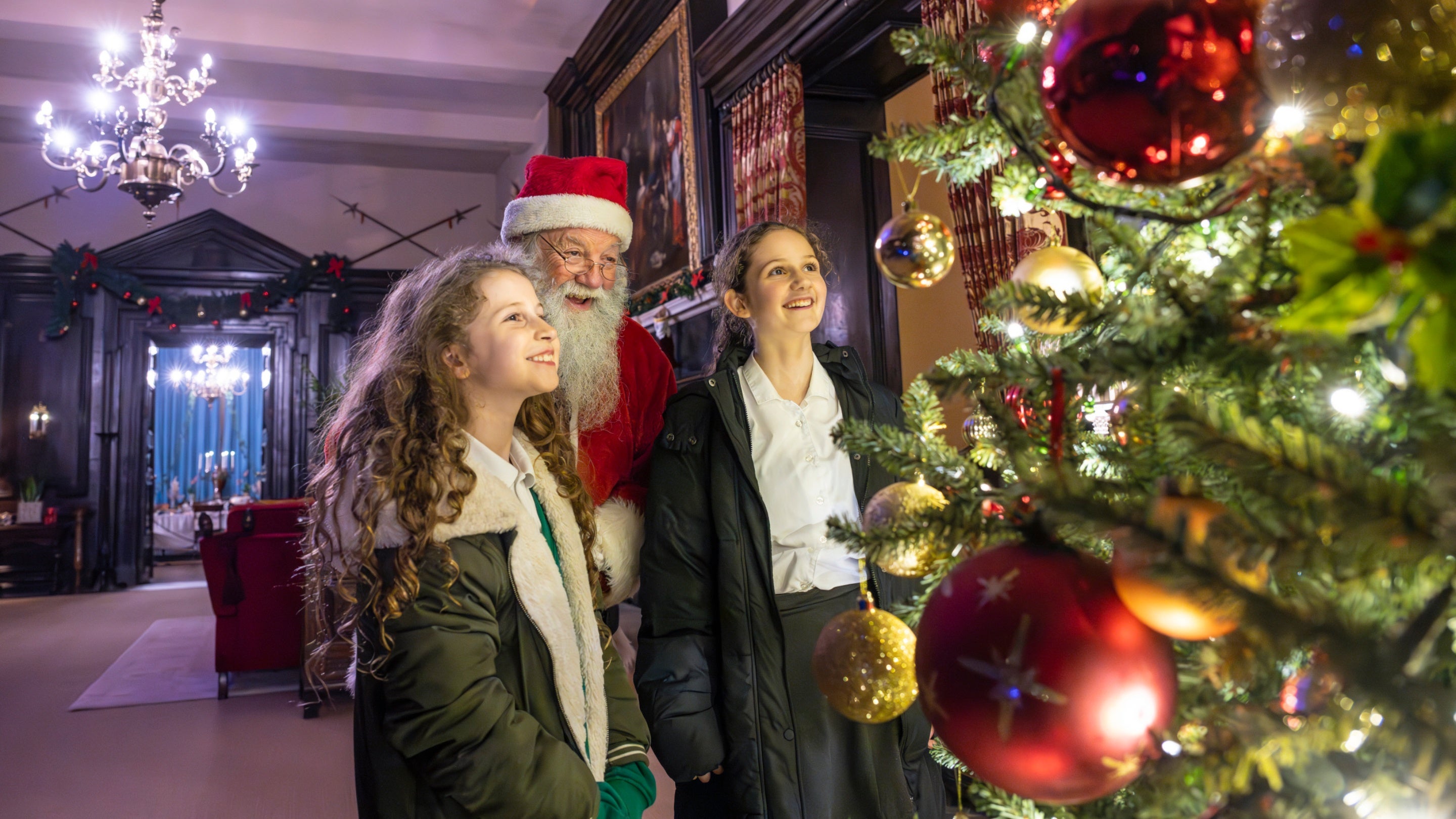 Two children and Father Christmas smiling and looking at a Christmas Tree decorated with red baubles and fairy lights in Tredegar House, Newport, Wales