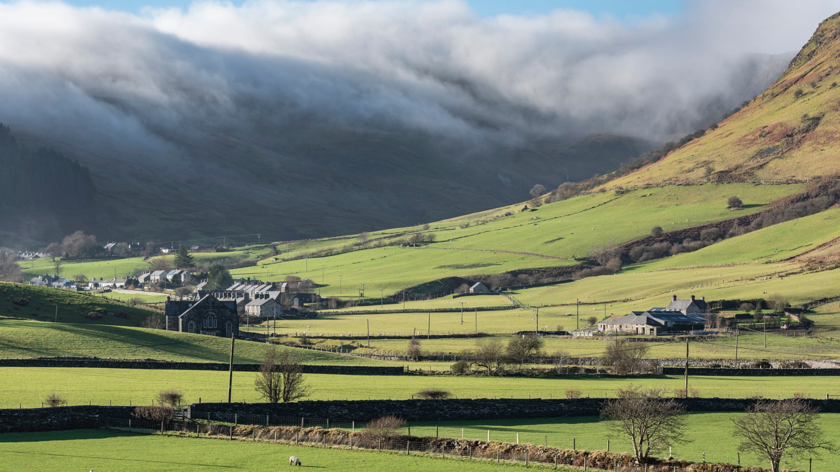 A sweeping view of green fields in the valley at Cwm Penmachno with some buildings visible in the valley and a sloping hill up the mountainside, with low-hanging clouds and a dark mountain in the background