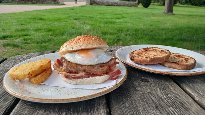A breakfast bap consisting of bacon, sausages and a fried egg in a brioche bap, with hash browns on the side. Available from the Barn Restaurant at Baddesley Clinton, Warwickshire.