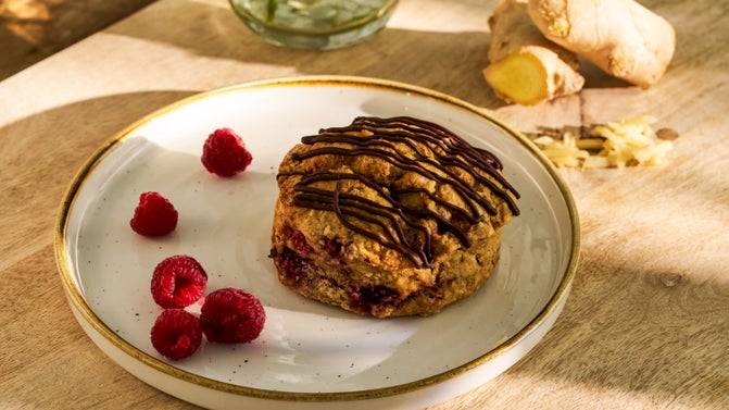A plate with some fresh raspberries and a delicious raspberry and ginger scone drizzled with chocolate sauce. In the background some pieces of raw and grated ginger.