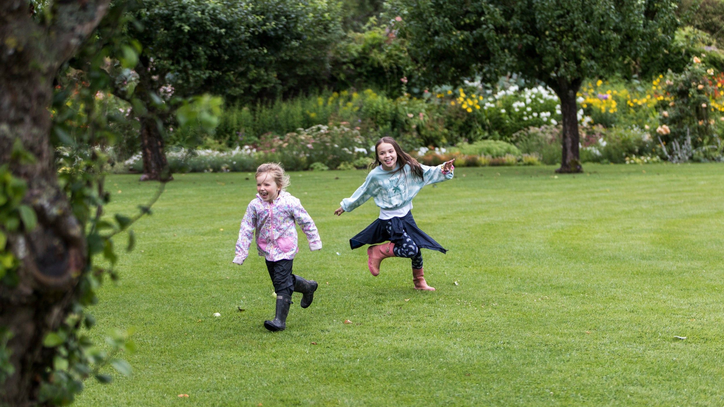 Two girls wearing wellies and raincoats running across the lawn in a walled garden with  trees and a herbaceous border in the background