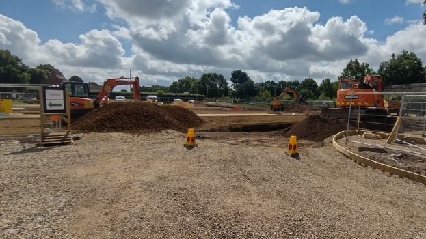 A view across the car park with mounds of earth and diggers either side, framed by herace fencing.