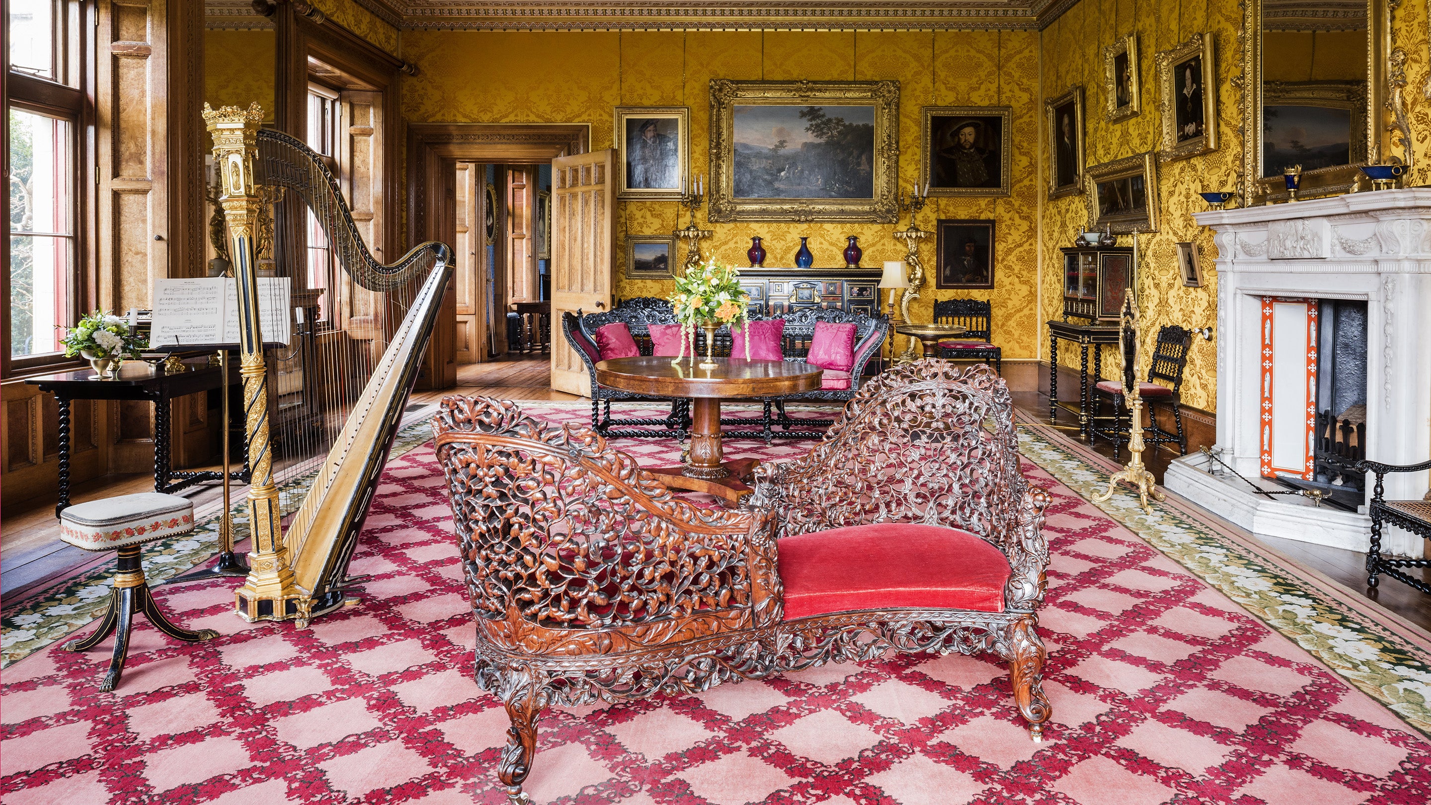 The drawing room has bright yellow, brocade wallpaper, a large pink and red carpet, the wall is adorned with paintings, there is a large table with chairs, a 'kissing' seat, and a harp.