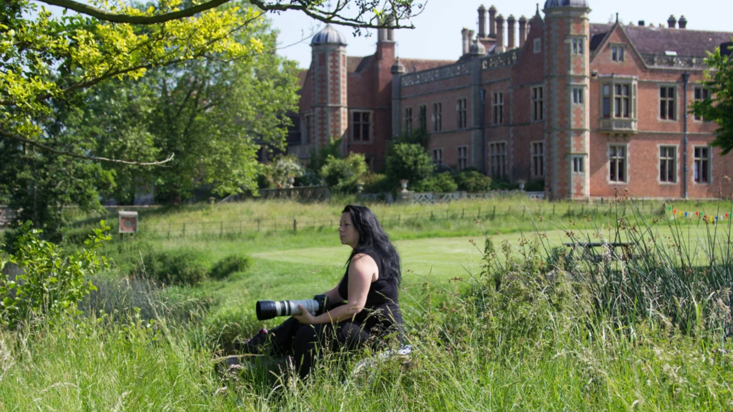 Jana has dark hair which is blowing slightly in the wind. She is holding a camera and is looking out across the river, surrounded by green grass with Charlecote house in the background.