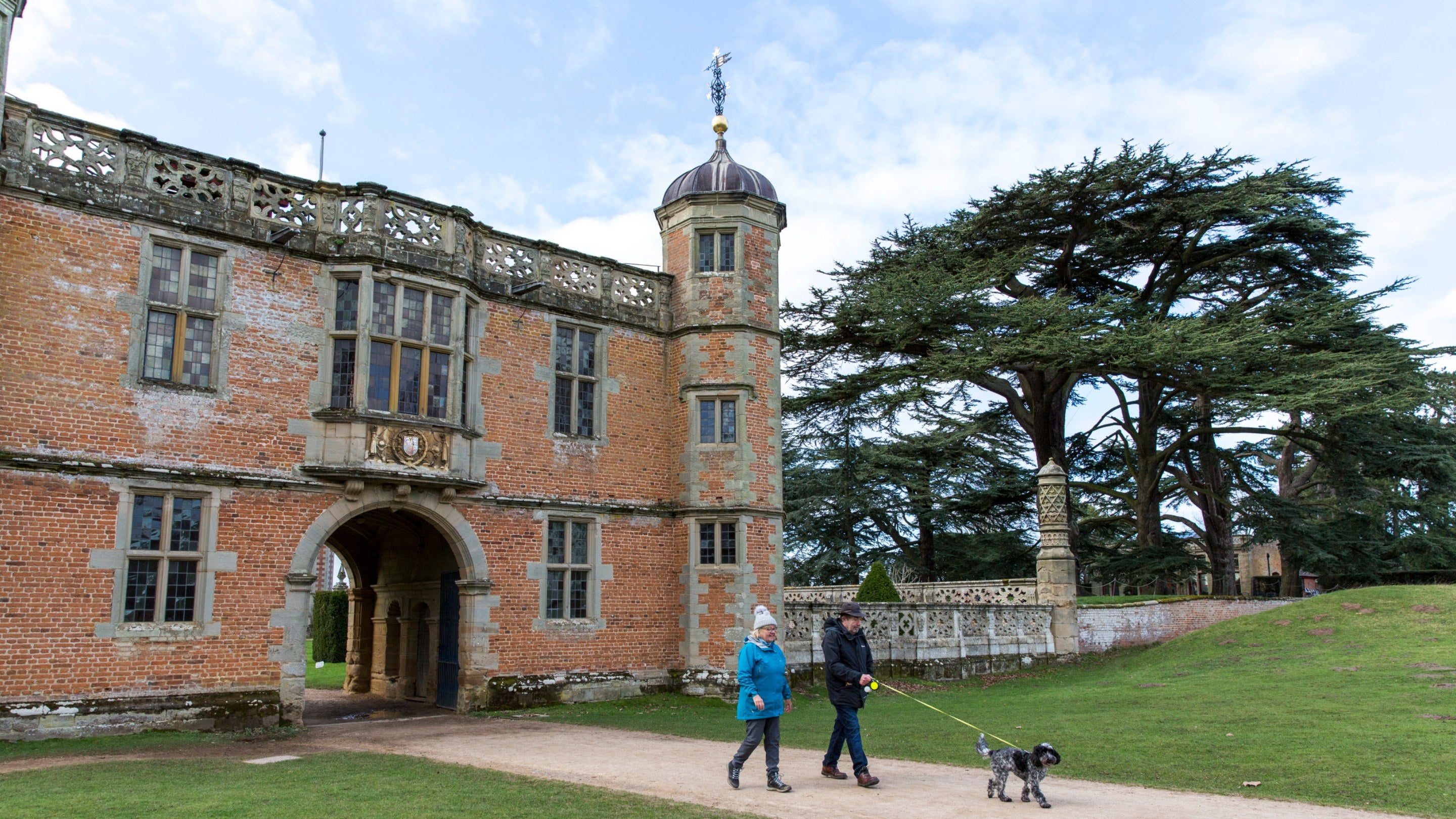 Two dog walkers with their dog on a lead walk away from the red brick gatehouse at Charlecote Park