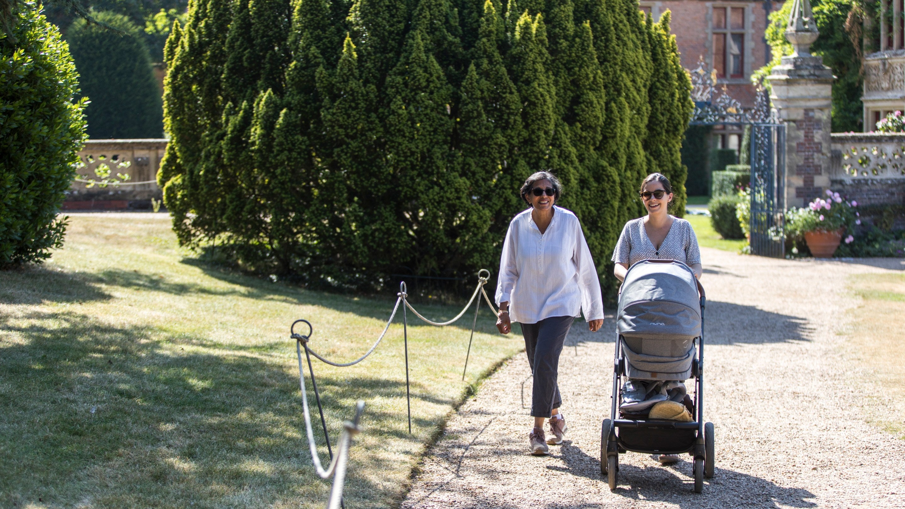 A multi generational family taking a leisurely stroll along a shady path in the gardens on a bright, hot sunny summer's day. A mature daughter is talking to her mother (grandmother) who is pushing a pram.